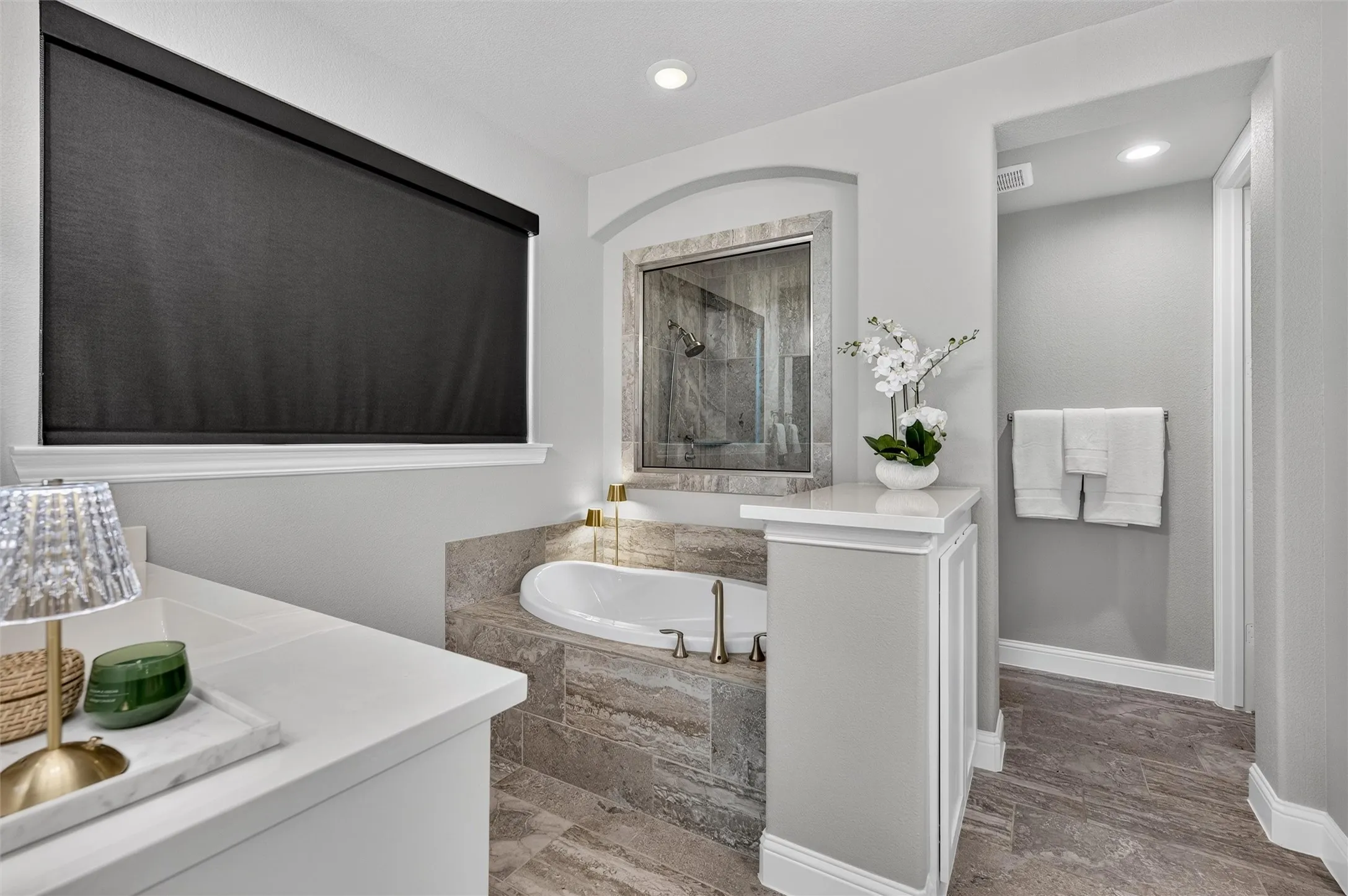 Soaking tub with stone surround, walk-in shower with glass enclosure, recessed lighting, and large window with roller shade