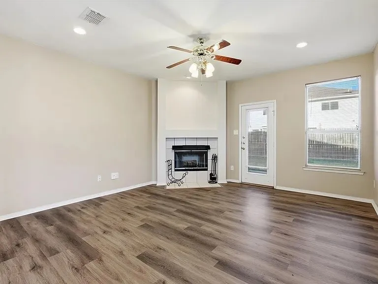Spacious room featuring wood-finish flooring, a corner fireplace with a white tile surround, a ceiling fan with integrated lighting, recessed lighting, and a glass-paneled exterior door