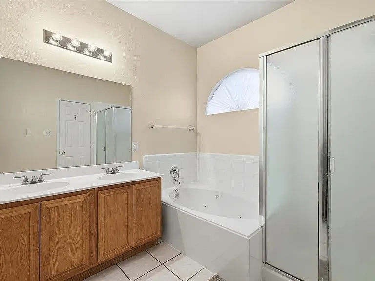 Bathroom featuring a double vanity with wood cabinetry, a large built-in mirror, an integrated soaking tub with white tile surround, a separate shower with frosted glass doors, and a decorative arched window