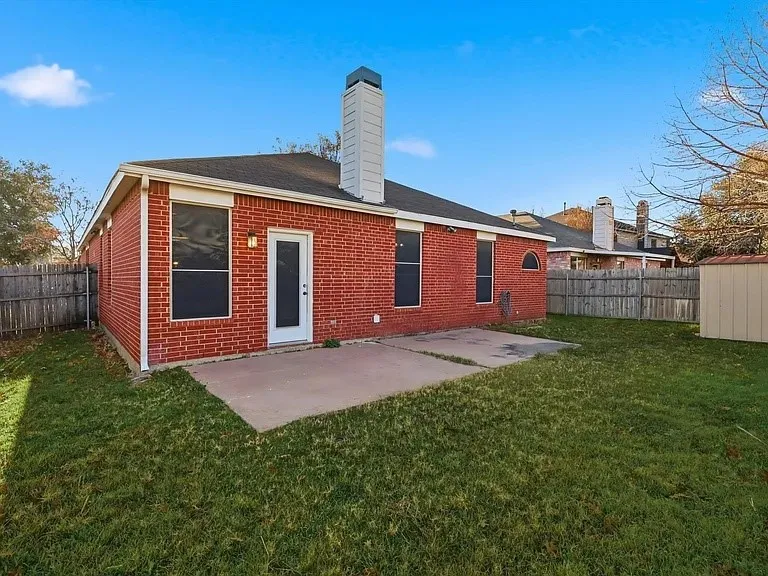 Red brick exterior with a white trim door and windows, a concrete patio, a wooden privacy fence, and a white chimney