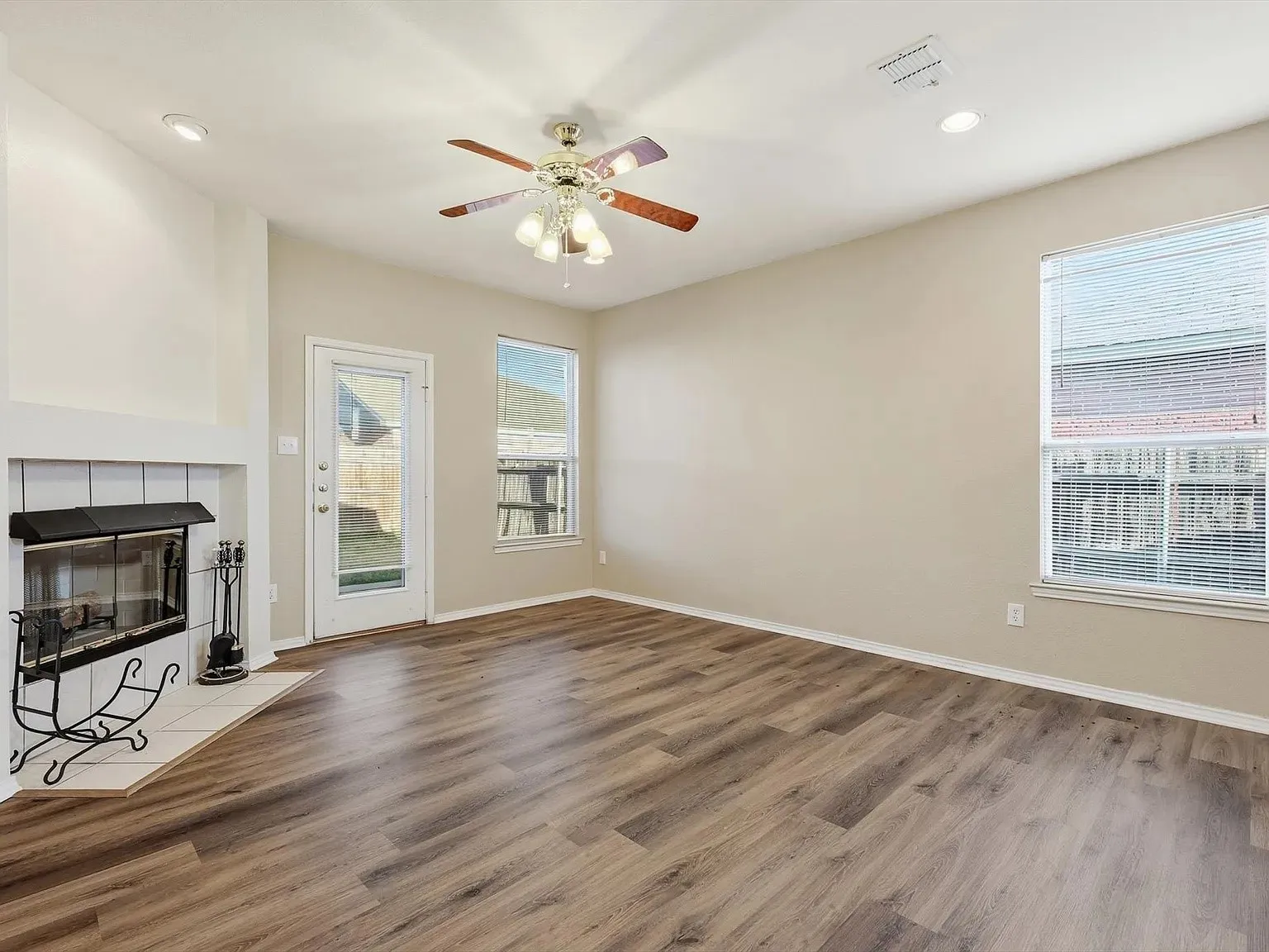 Spacious room with wood-finish flooring, a white mantel fireplace with hearth, multiple windows, and a ceiling fan with integrated lighting