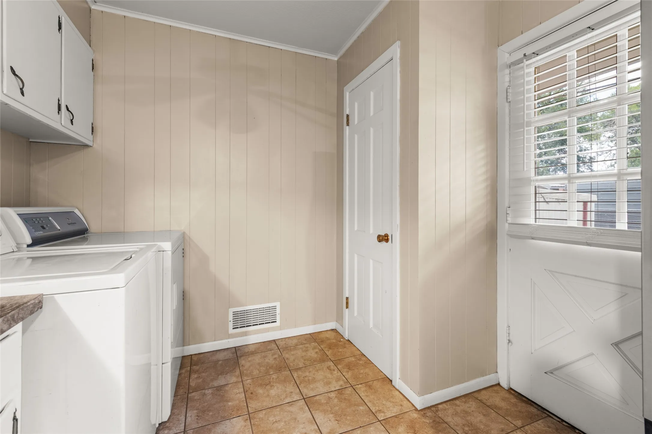 Utility space with paneling walls, tiled flooring, white cabinetry, and a split exterior door with window blinds
