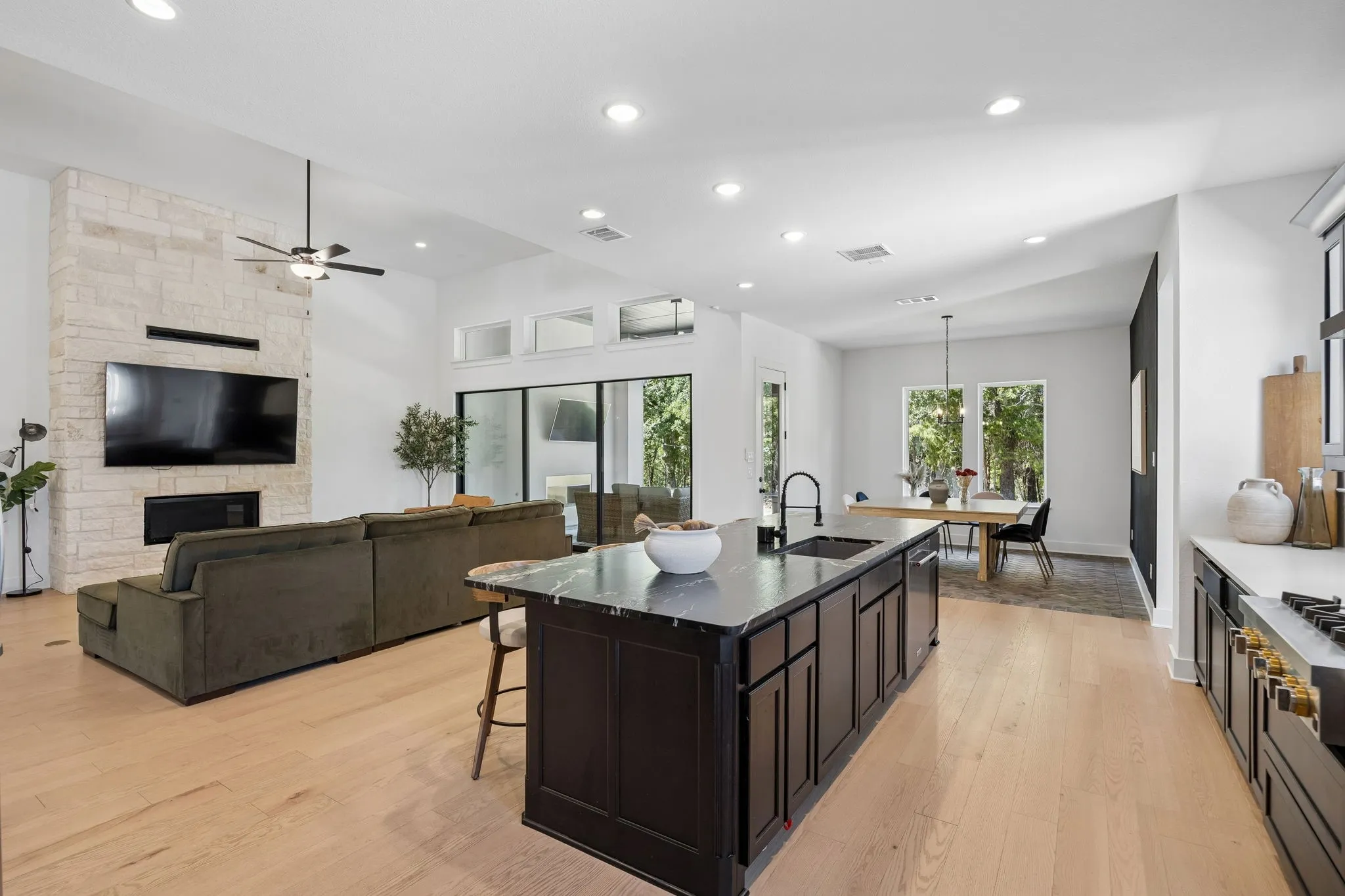Open-concept living space featuring light wood-finish flooring, a stacked stone fireplace surround, a kitchen island with dark cabinetry, ample recessed lighting, and large windows
