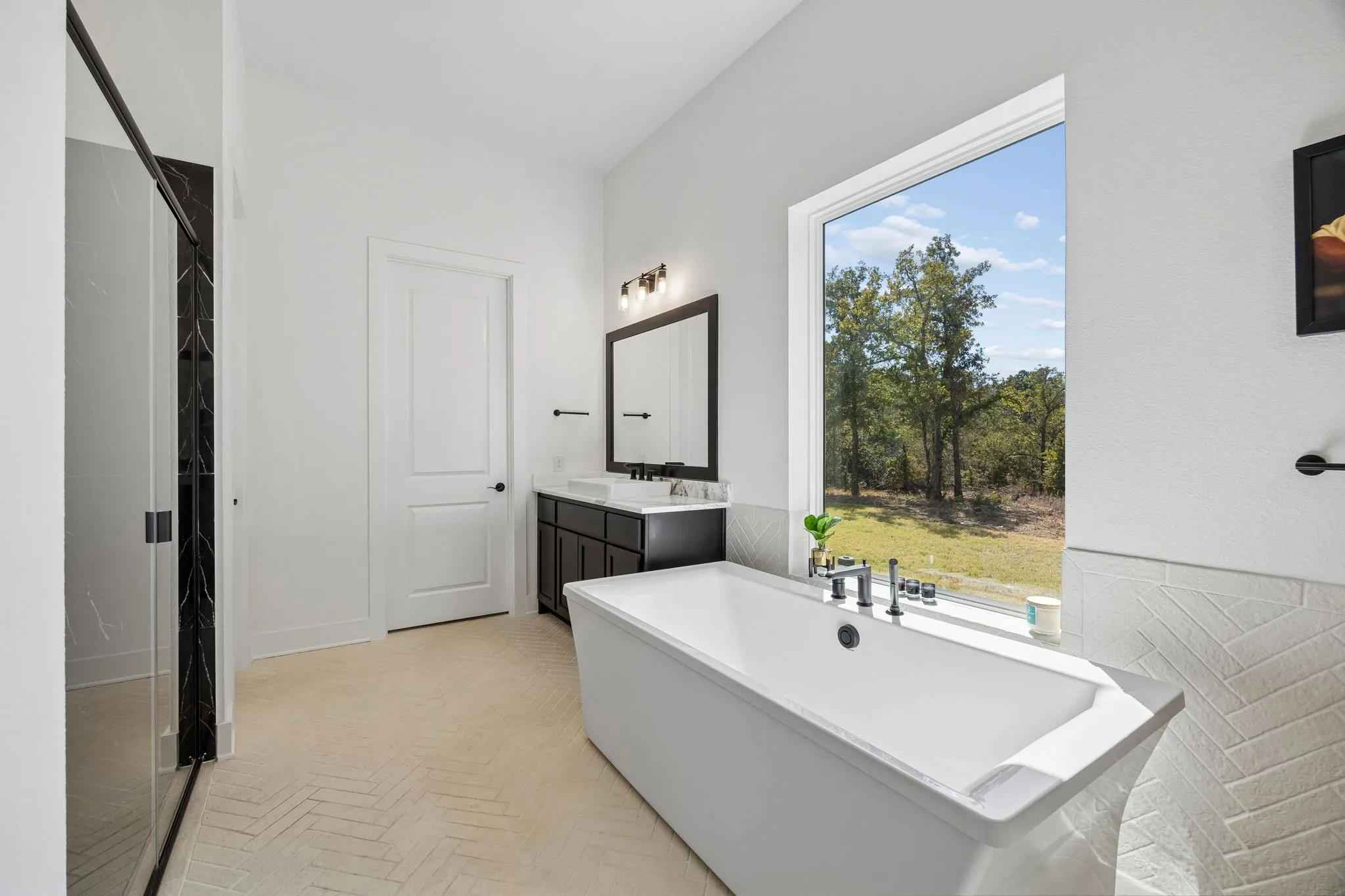 Freestanding soaking tub, large window, herringbone tile flooring, dark wood-finish vanity with dual sinks, and a glass-enclosed shower
