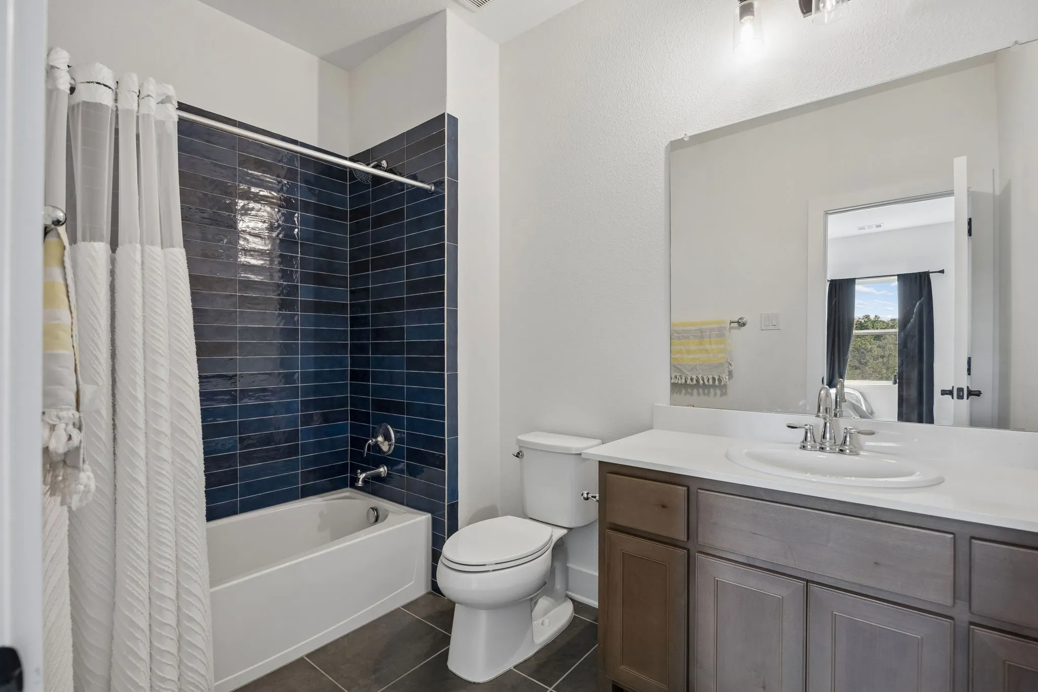 Bathroom featuring a tub with a deep blue tile surround, a single vanity with an integrated sink, a large wall-mounted mirror, and dark tile flooring