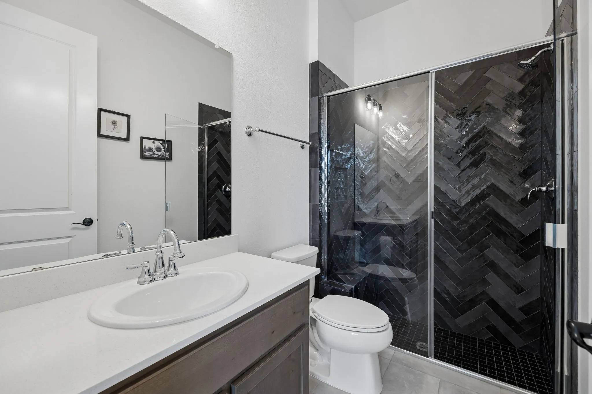 Vanity with an integrated sink, shower with dark herringbone tile surround, glass shower enclosure, and chrome fixtures