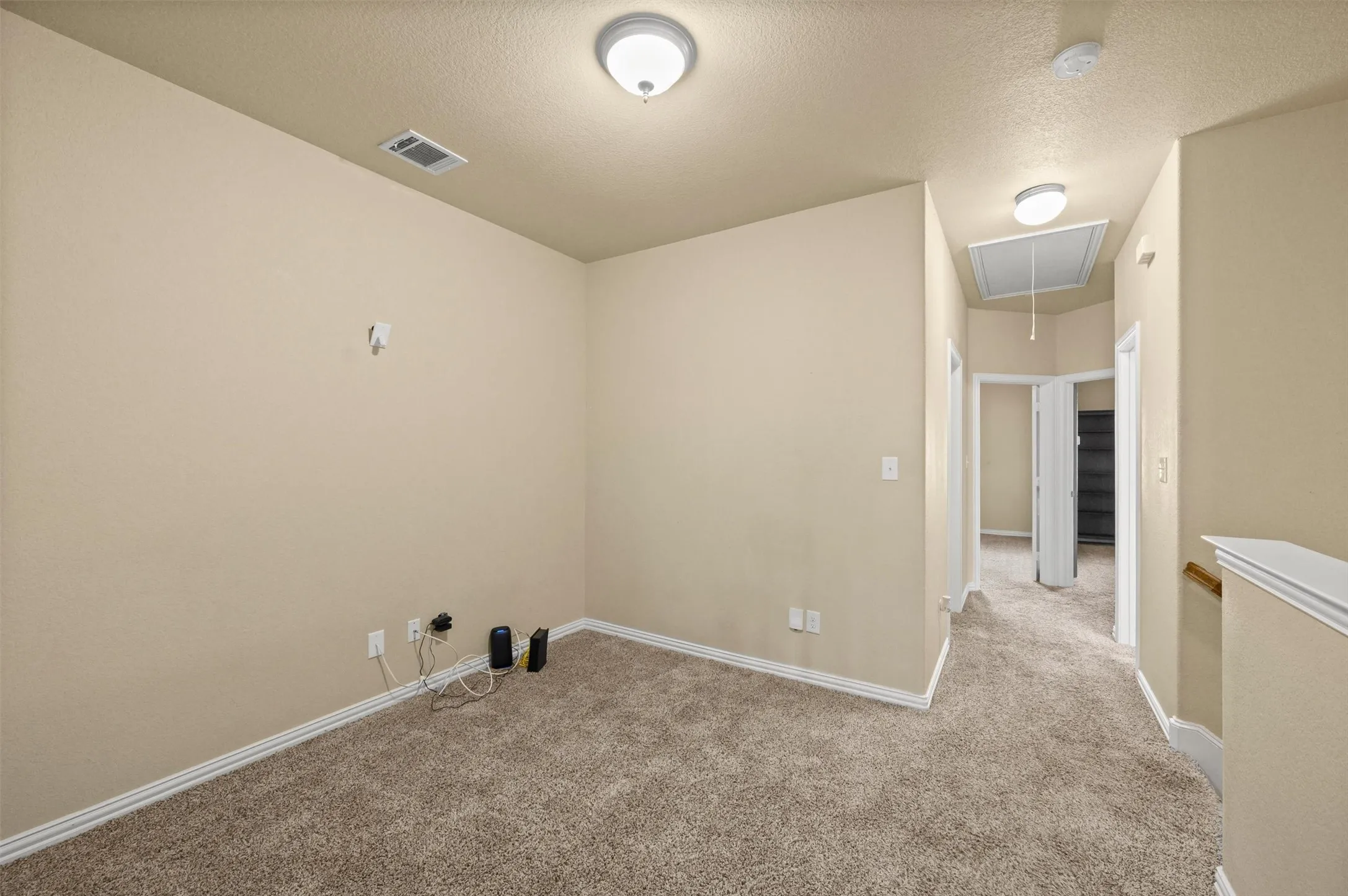 Carpeted room with neutral wall color, ceiling-mounted light fixtures, and a hallway with an attic access panel