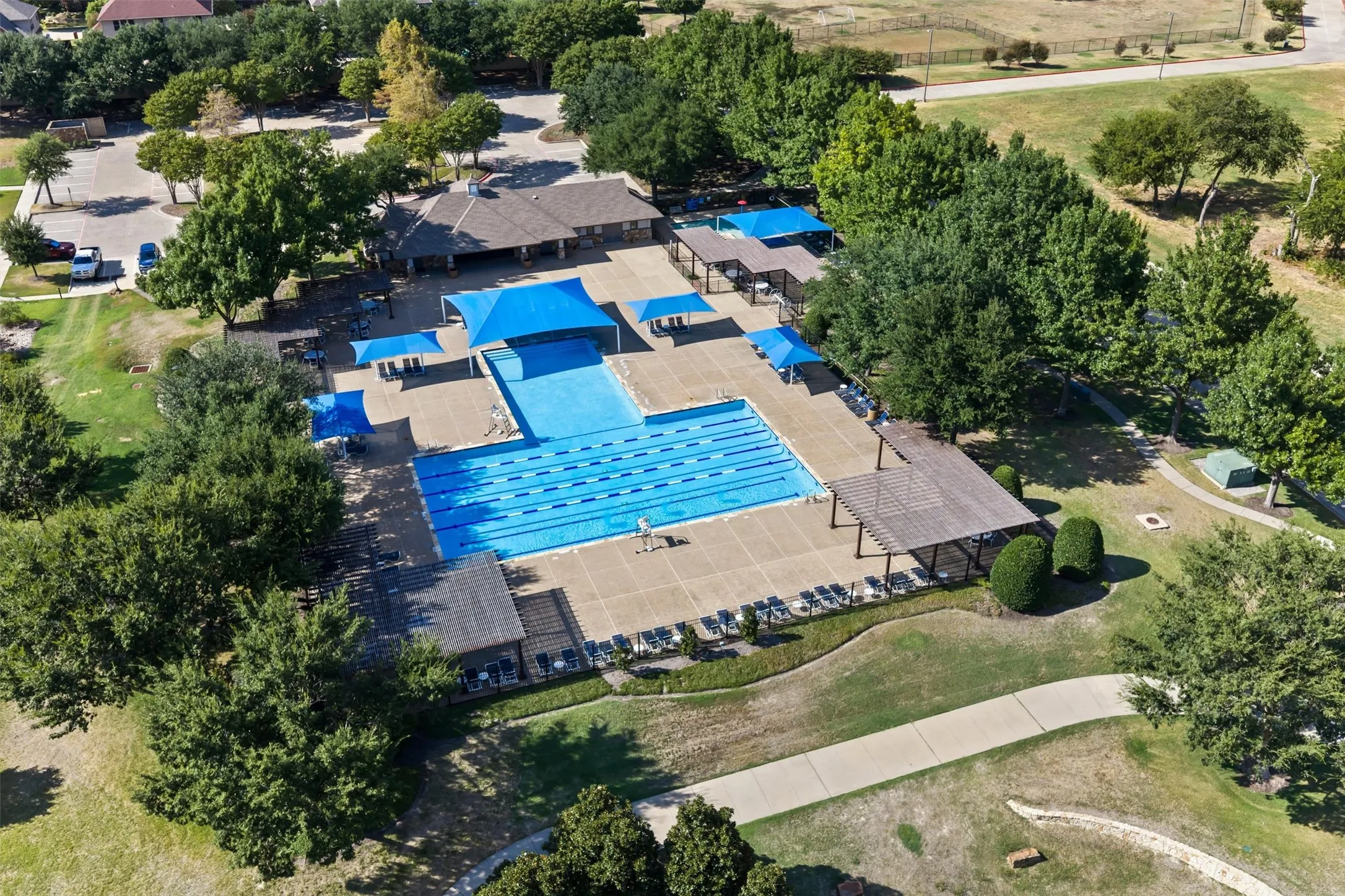 Community pool with lap lanes, adjacent covered seating areas, surrounding hardscaping, and a nearby community building with an adjacent parking area