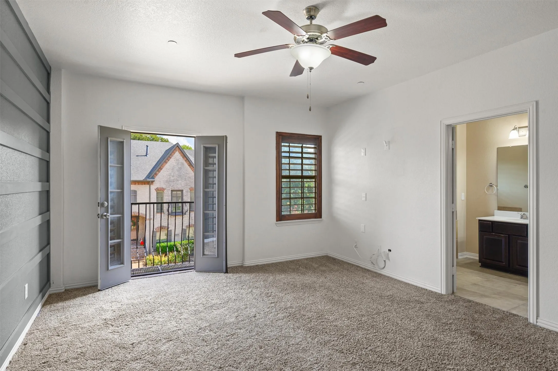 Carpeted flooring, ceiling fan with light fixture, French doors opening to a balcony with an iron railing, and a window with plantation shutters