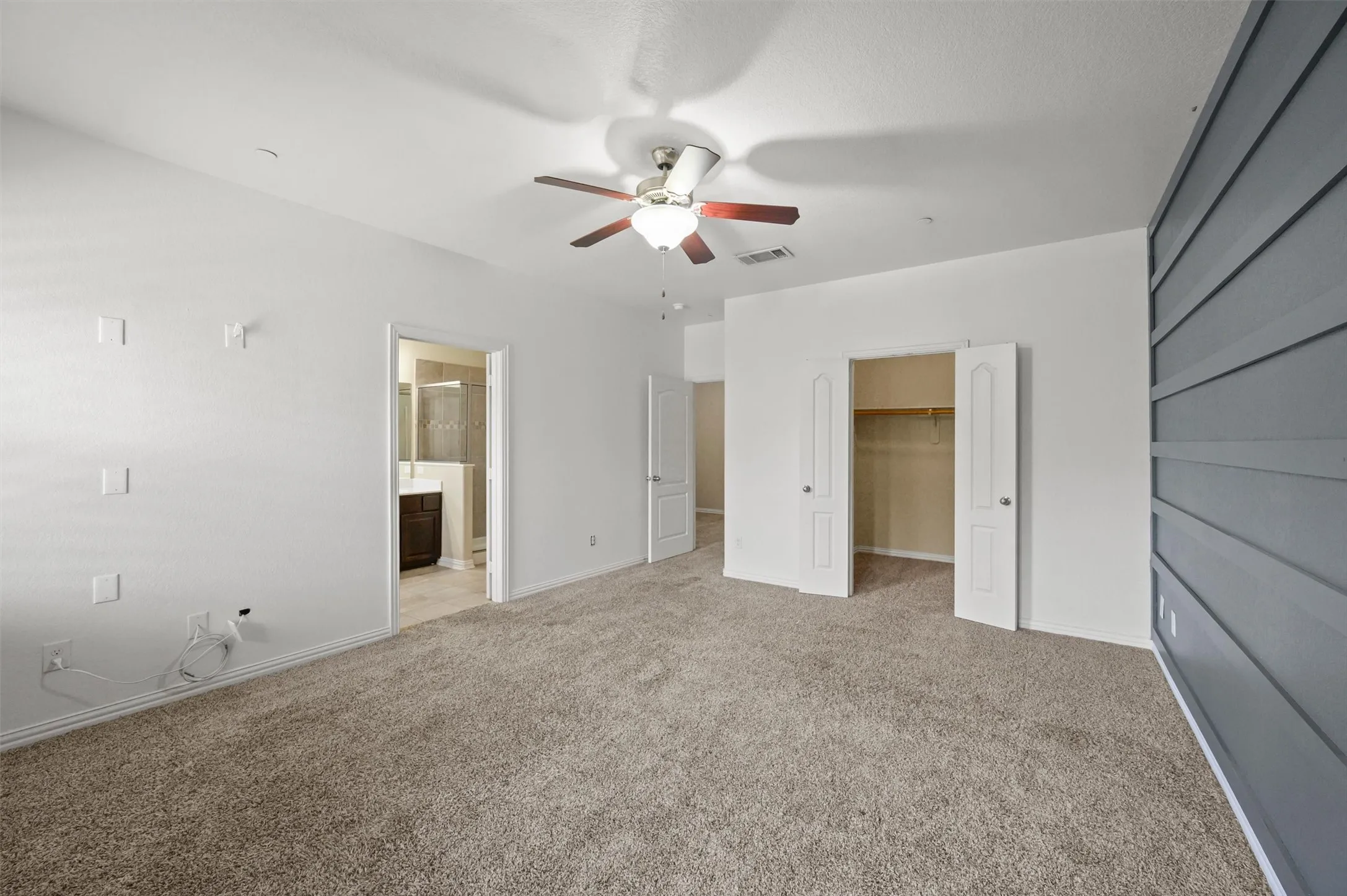 Carpeted flooring, ceiling fan with light fixture, built-in closet with double bifold doors, accent wall with horizontal paneling, and an adjacent bathroom with a dark wood-finish vanity