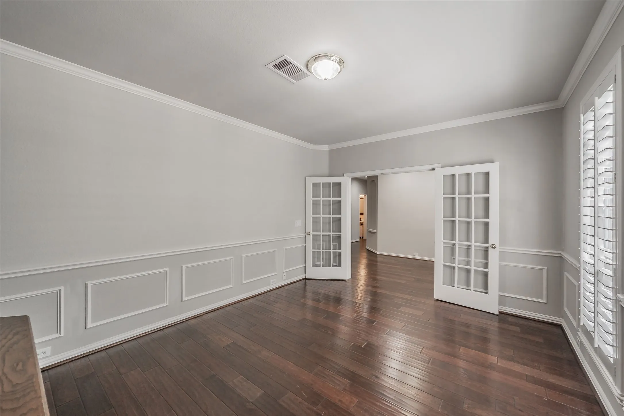 Dark wood-finish flooring, wainscoting, crown molding, French doors with grid panes, and plantation shutters