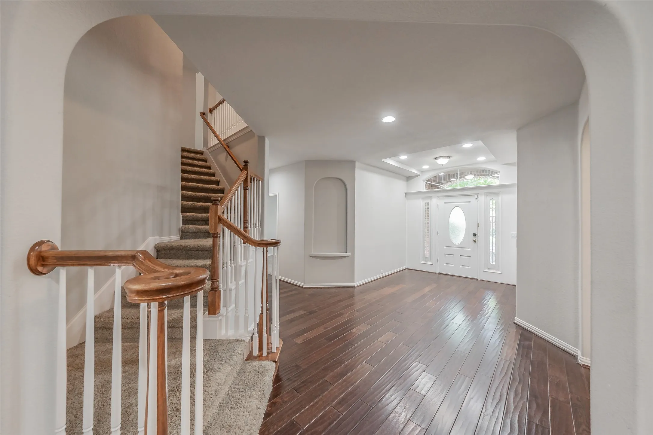 Arched entryway featuring wood-finish flooring, a carpeted staircase with wood handrails, a white paneled front door with an oval glass insert, and recessed ceiling lighting
