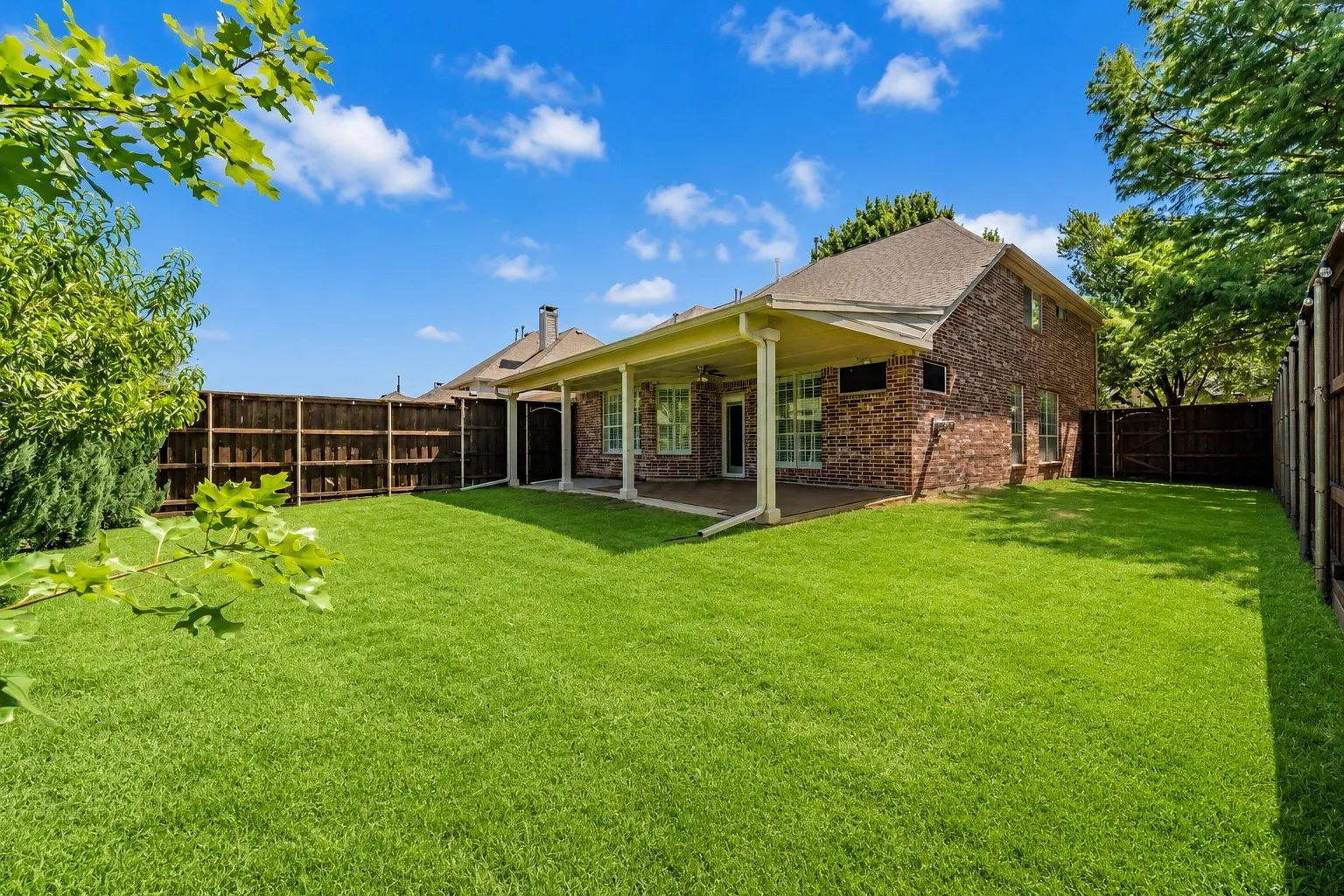 Expansive green lawn with a covered patio featuring support columns, a brick exterior, and wood fencing