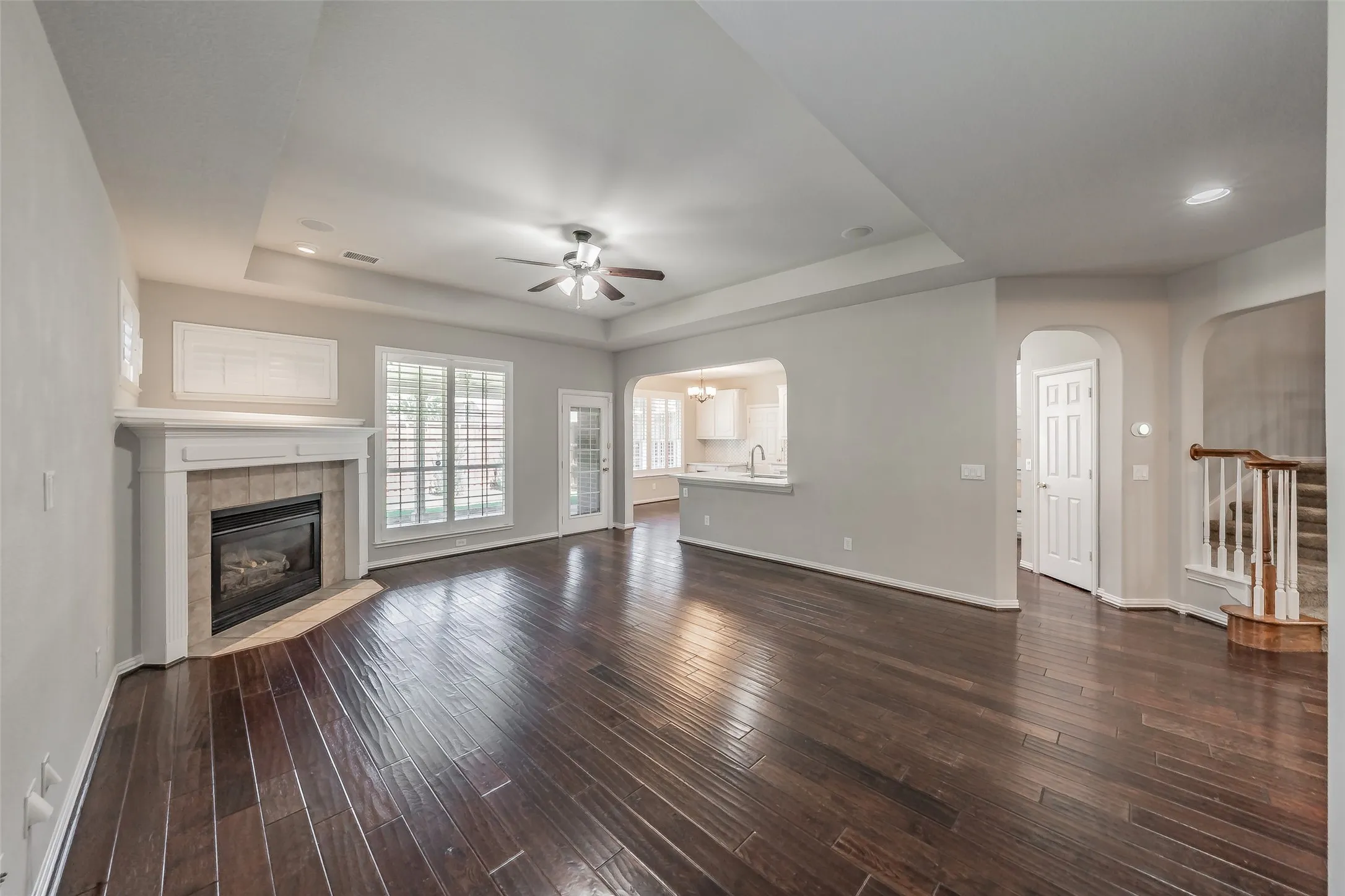 Dark wood-finish flooring throughout, tray ceiling with a ceiling fan, fireplace with a tile surround and white mantel, recessed lighting, and an arched doorway
