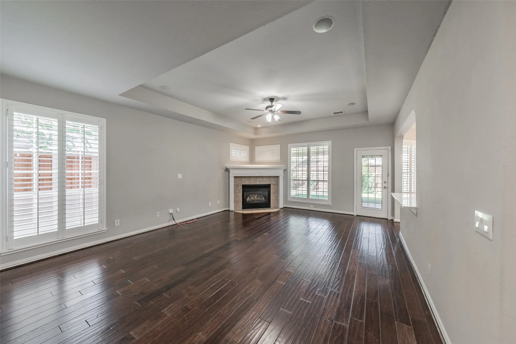 Dark wood-finish flooring, corner fireplace with mantel, tray ceiling, ceiling fan, and multiple windows with plantation shutters