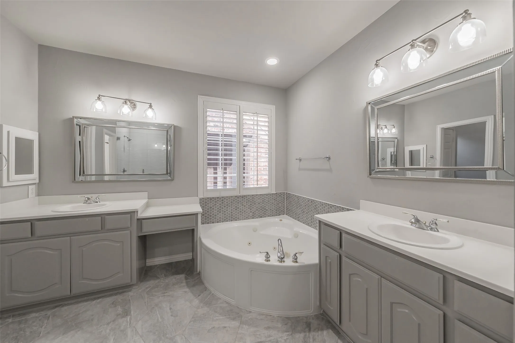 Dual vanities with undermount sinks and light gray cabinetry, a corner built-in soaking tub with mosaic tile surround, and plantation shutters