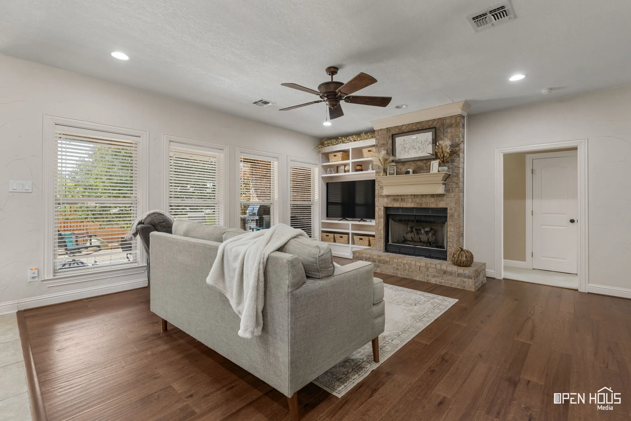 Wood-finish flooring throughout, brick fireplace with a mantle, built-in shelving, ceiling fan, and recessed lighting