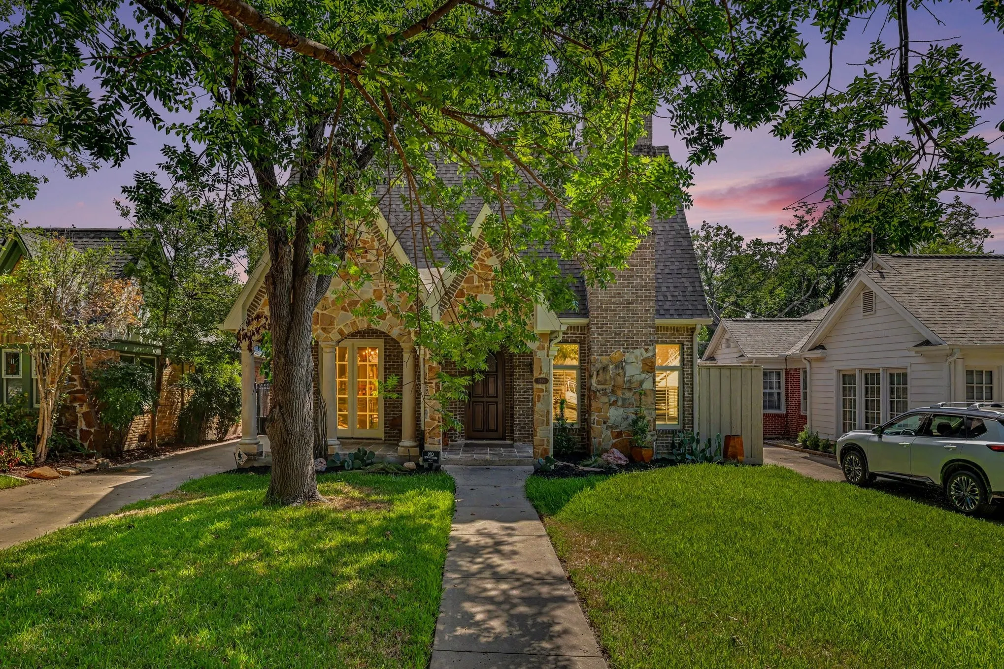 Stone and brick facade, gabled roofline, arched entry, mature tree, and concrete pathway