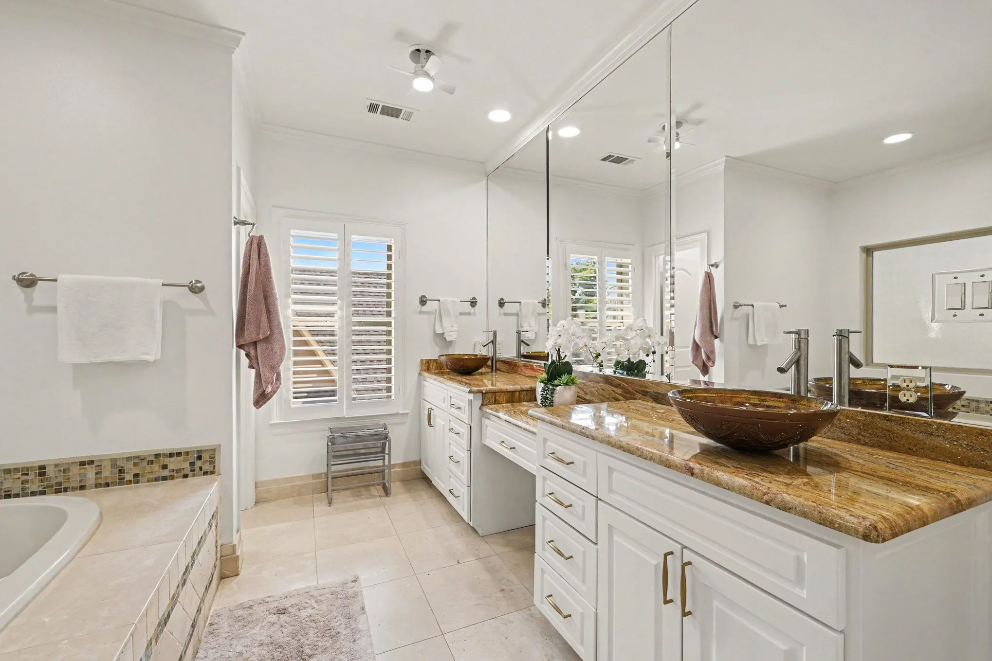 Dual vessel sinks set on a stone countertop with white cabinetry, extensive wall-mounted mirrors, and ceiling fan