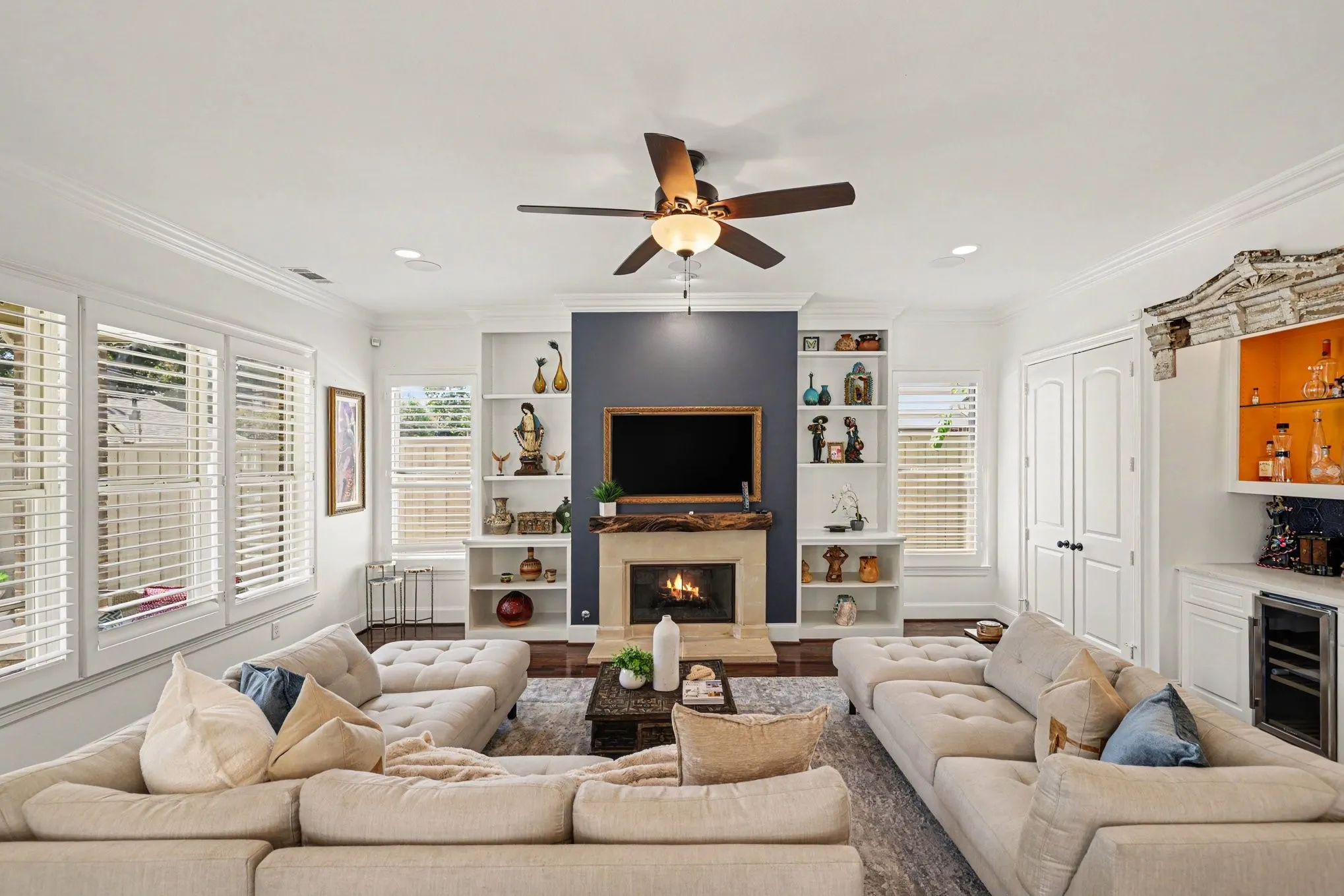Living area featuring a fireplace with a mantel, built-in shelving, wood-finish flooring, a ceiling fan, and plantation shutters