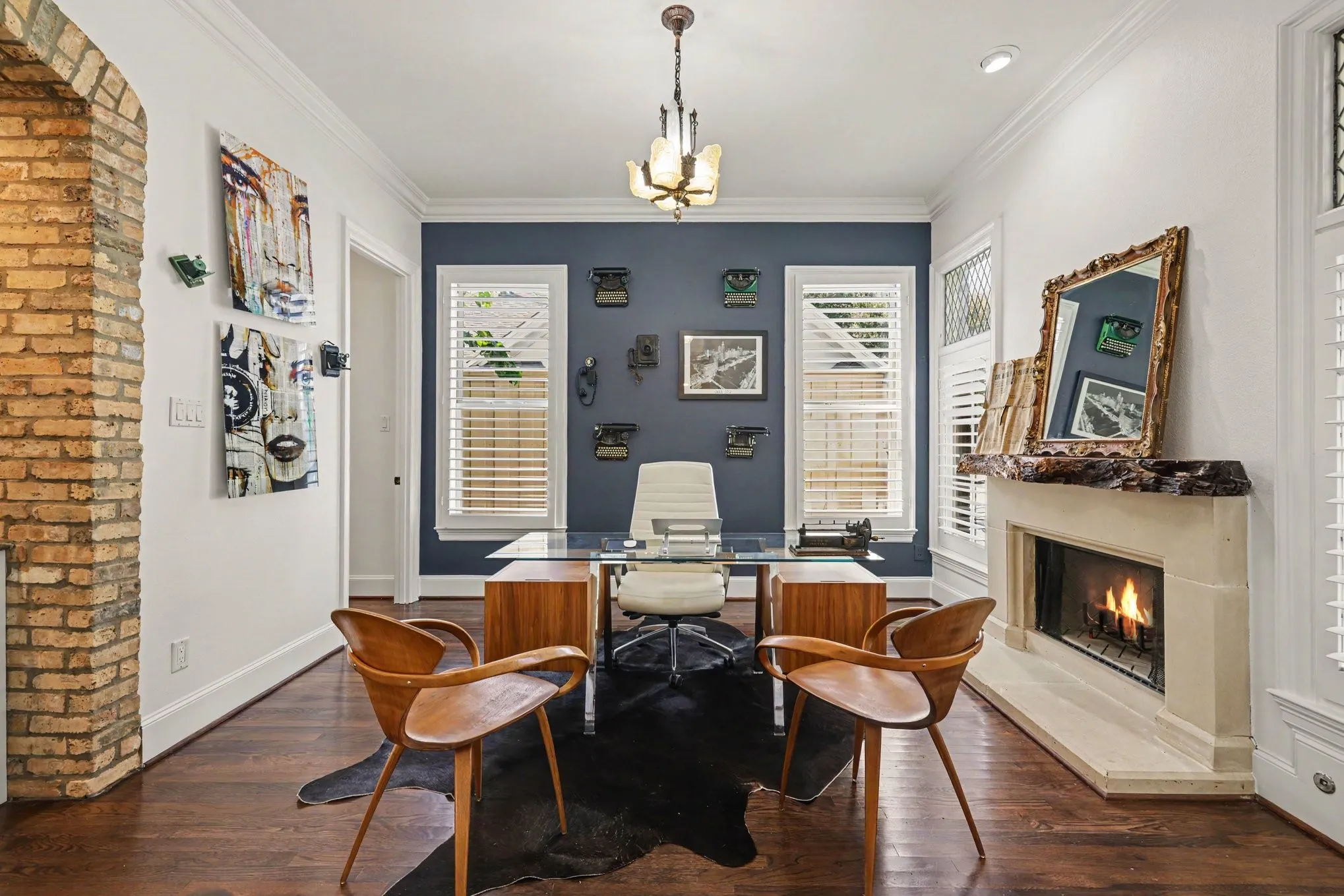 Hardwood flooring throughout, featuring a brick archway, a built-in fireplace with a stone surround, windows with louvered shutters, and a ceiling-mounted light fixture