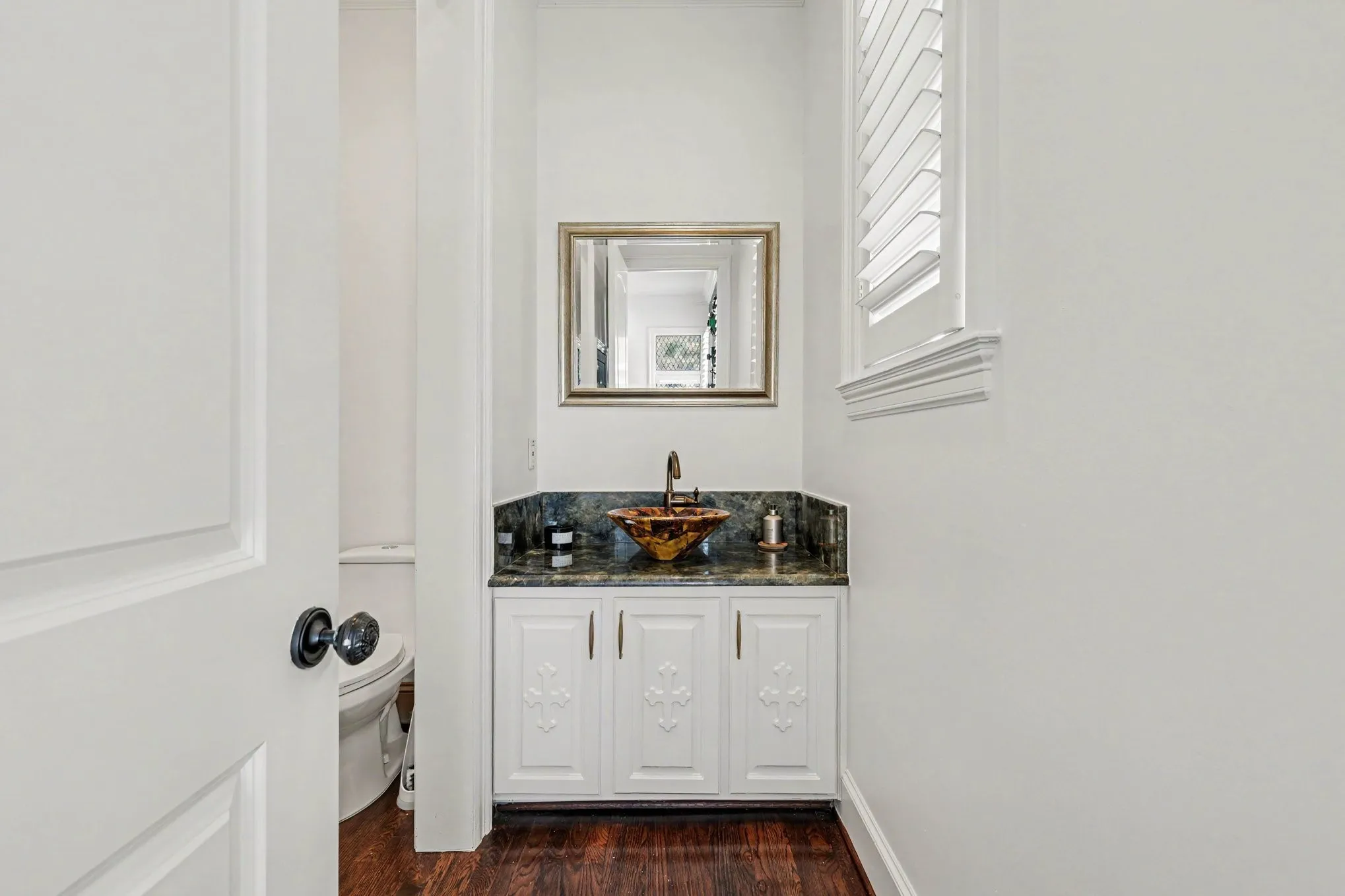 Powder room featuring a decorative vessel sink, dark stone countertop, white cabinetry with decorative panels, wood-finish flooring, and window with plantation shutters