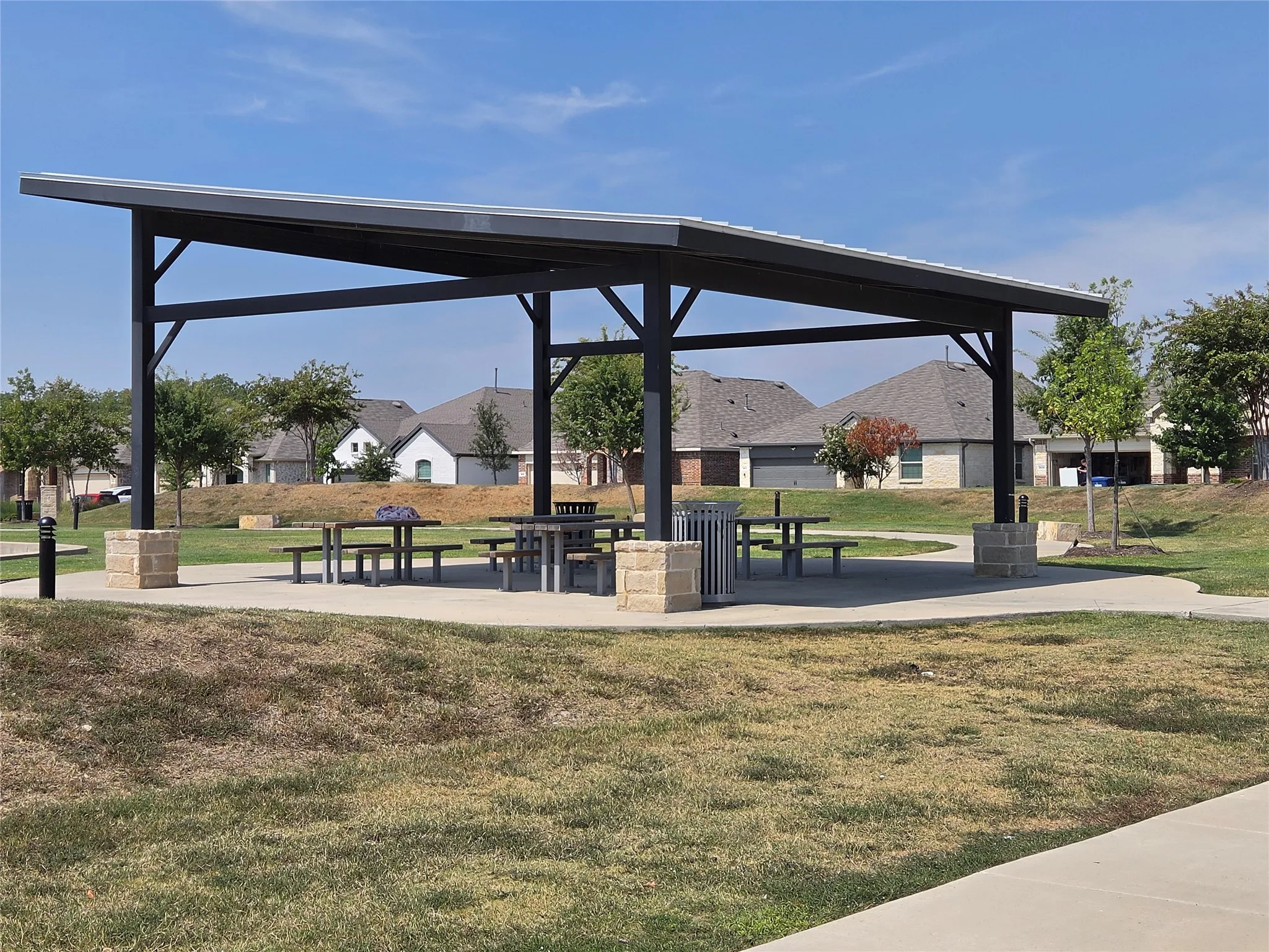 Expansive park pavilion with a slanted metal roof, multiple picnic tables, and a concrete pad