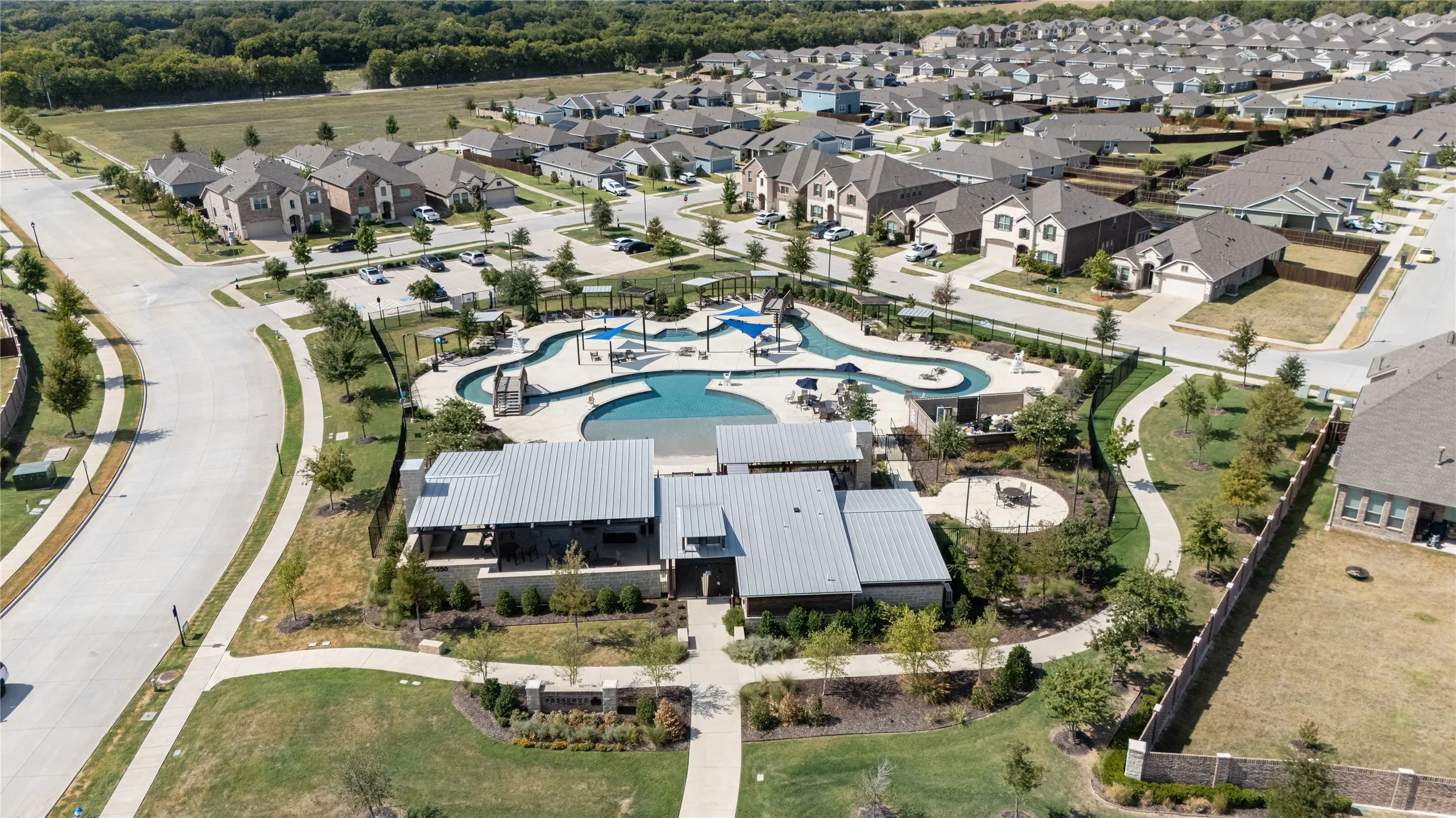 Community pool, adjacent water play area, and recreation building with covered outdoor spaces, surrounded by residential properties and green spaces