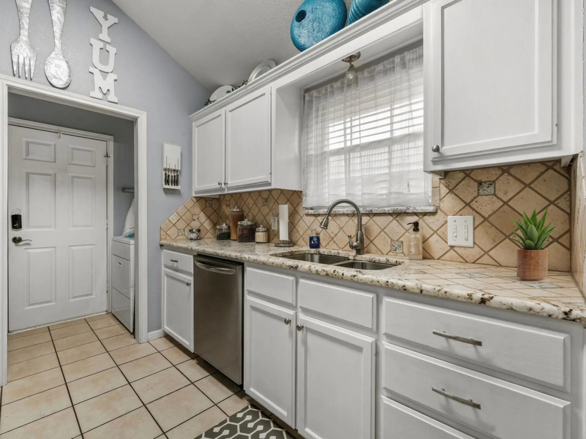 White cabinetry with brushed nickel hardware, light-toned tiled backsplash, granite-style countertops, stainless steel undermount double sink, and tiled flooring