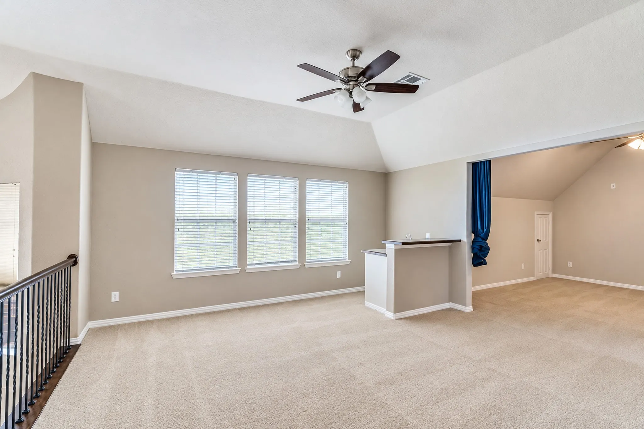 Spacious carpeted area featuring a built-in bar, multiple windows with blinds, a ceiling fan, and a wood railing with black spindles