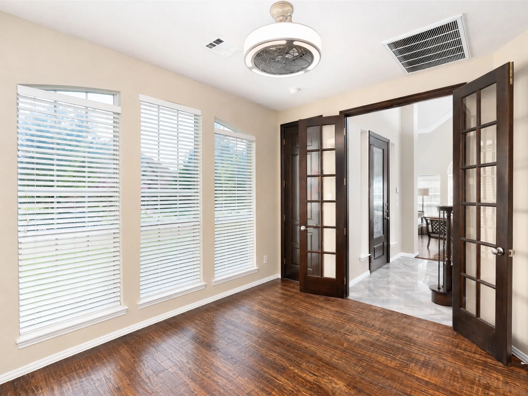 Wood-finish flooring, three tall windows with blinds, dual French doors with glass panels, and a ceiling-mounted fan fixture