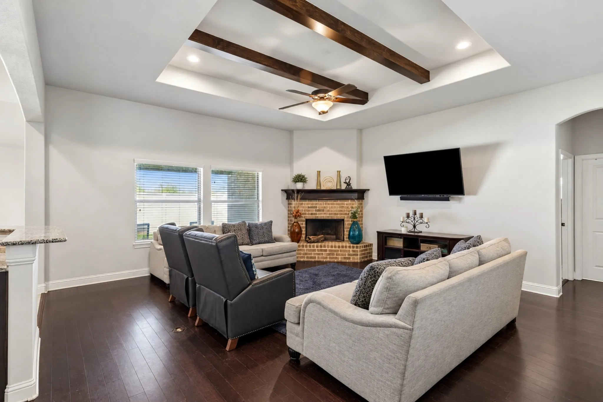 Tray ceiling with exposed wood beams, ceiling fan, dark wood-finish flooring, brick-surround fireplace with wood mantel, and arched doorway