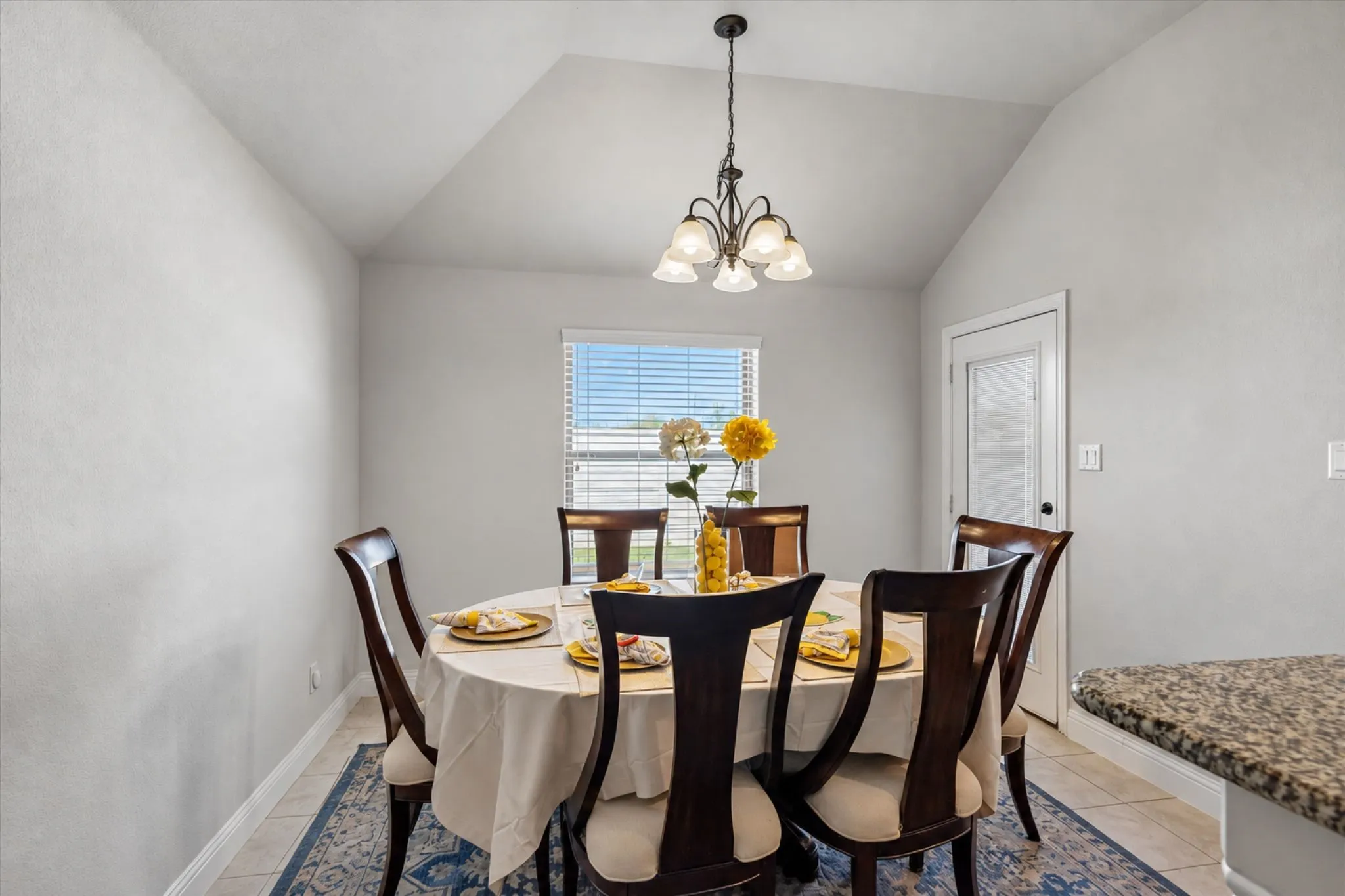 Vaulted ceiling, ceiling-mounted chandelier, window with blinds, tile flooring, and a glass-paneled door