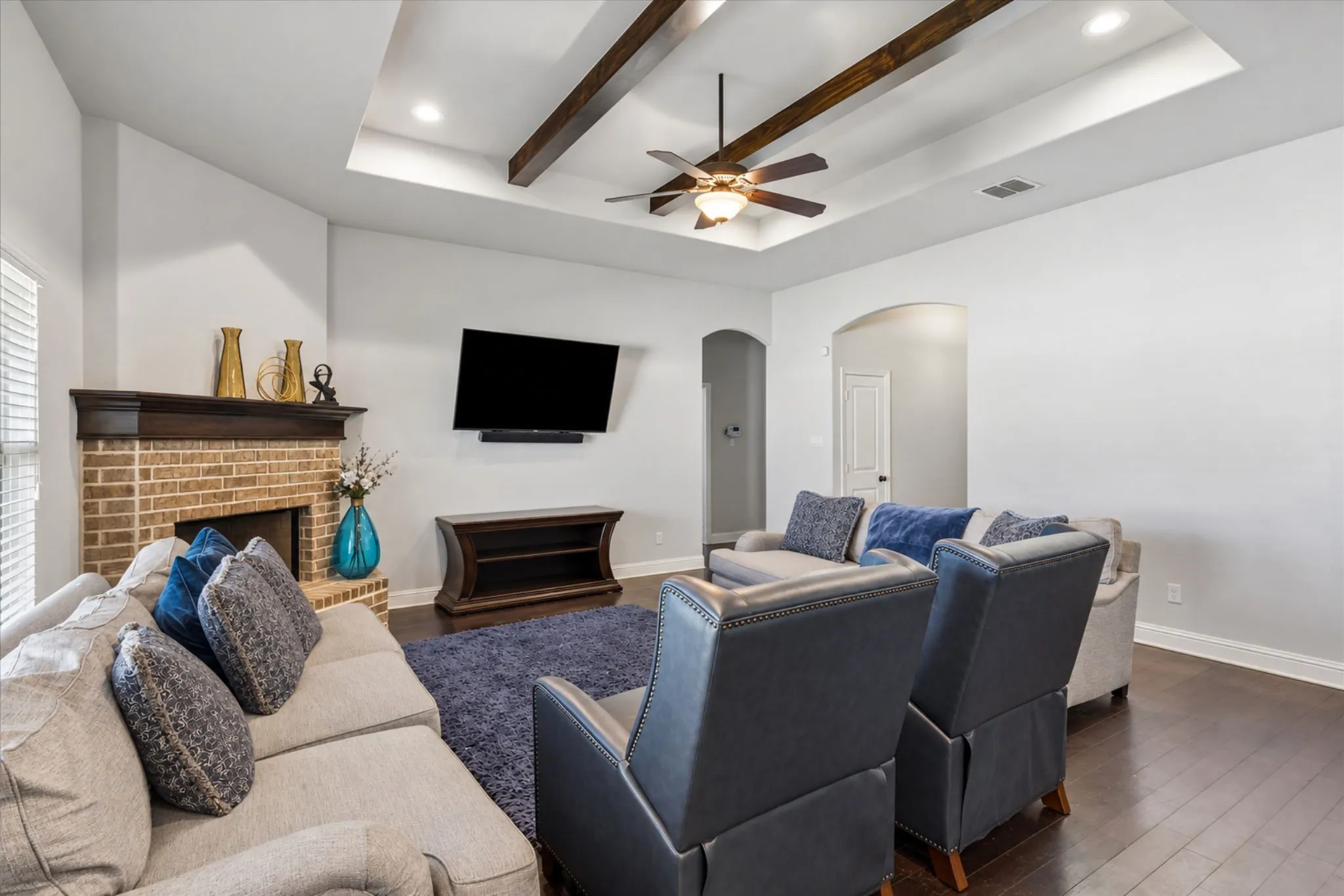 Tray ceiling with exposed wood beams, ceiling fan, recessed lighting, brick fireplace with wood mantel, and dark wood-finish flooring