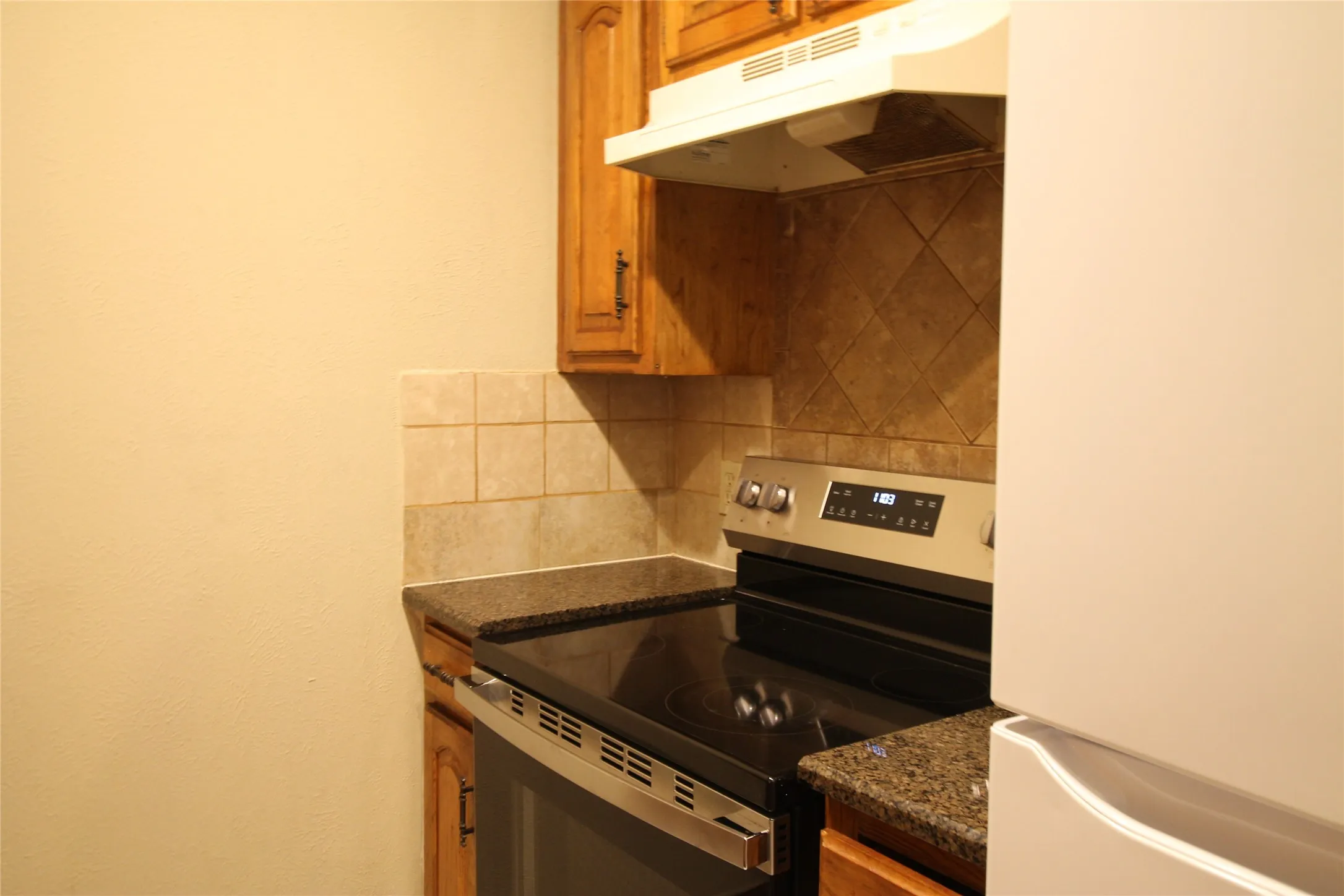 Stainless steel range, speckled countertops, wood-finish cabinetry, tiled backsplash, and range hood