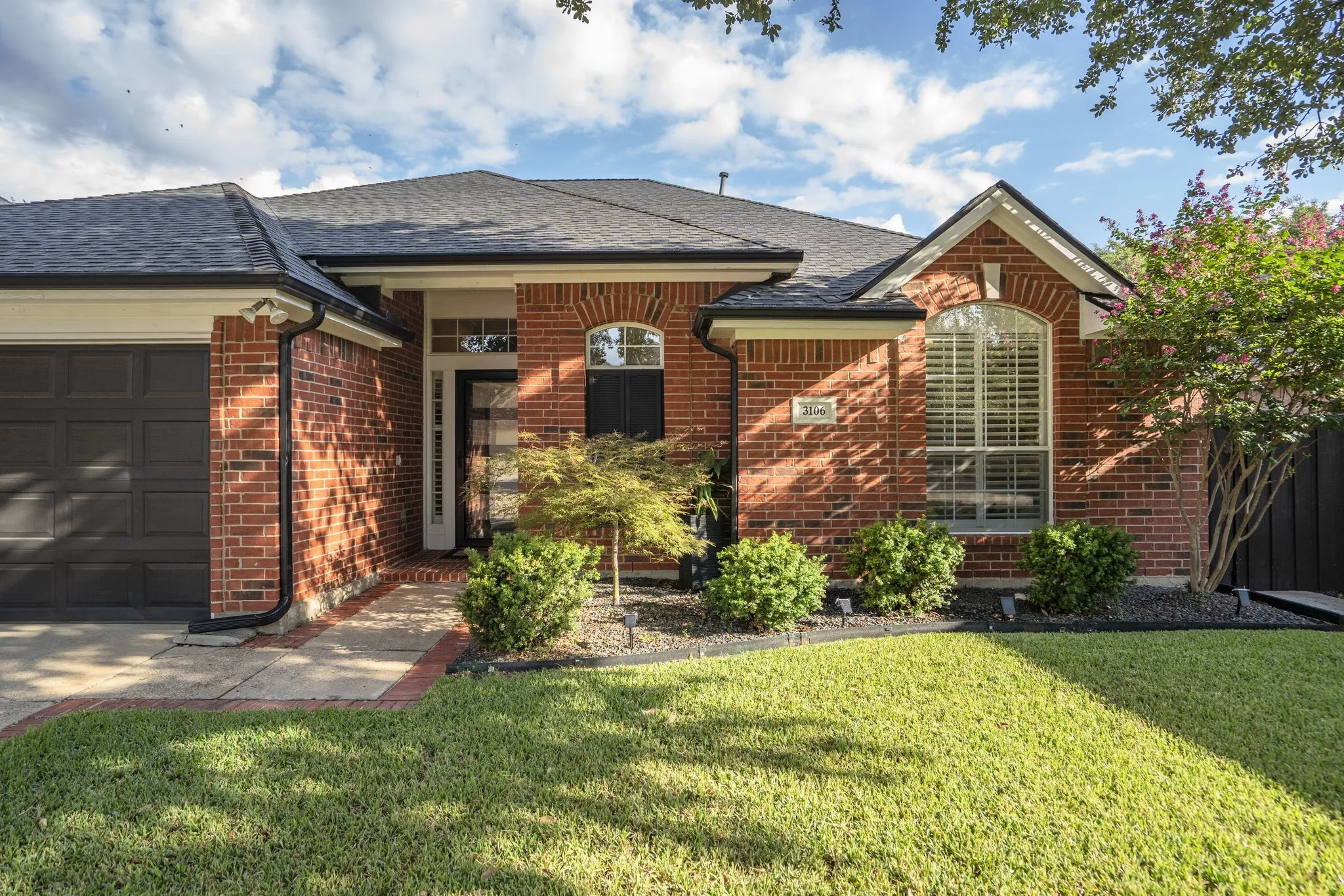 Red brick exterior with a dark shingle roof, a two-car garage, and an arched window with shutters