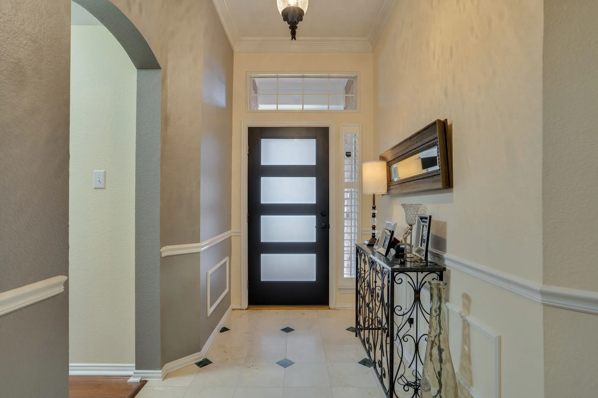 Entryway featuring a black door with frosted glass panels, transom window, sidelight with plantation shutters, tile flooring with diamond accents, and an arched doorway