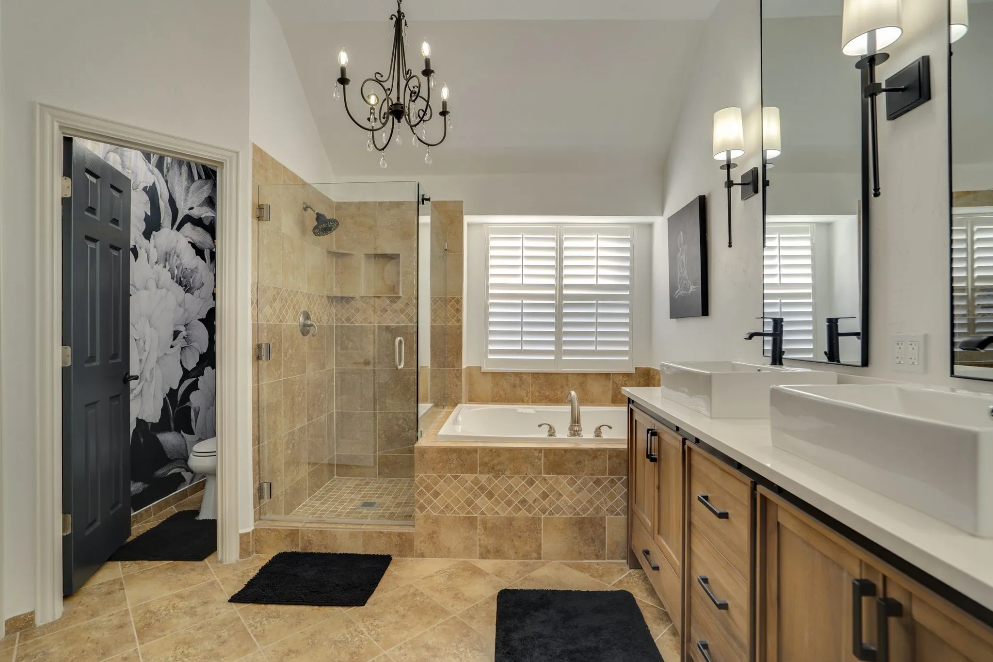 Dual vessel sinks with wall-mounted mirrors, a walk-in shower with glass enclosure, a built-in bathtub with tiled surround, plantation shutters, and a chandelier