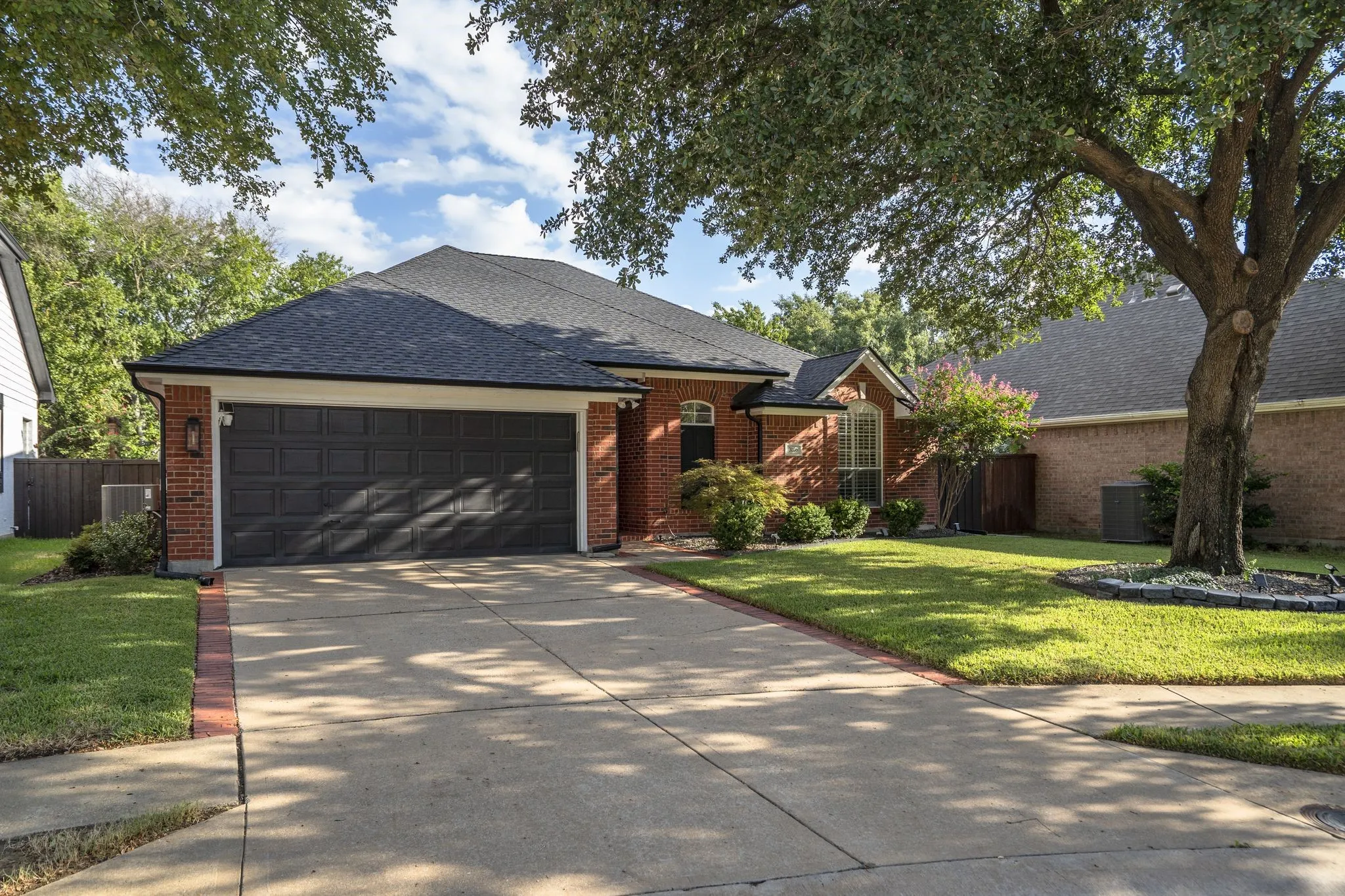 Red brick exterior, black asphalt shingle roof, two-car garage, concrete driveway, and established front landscaping