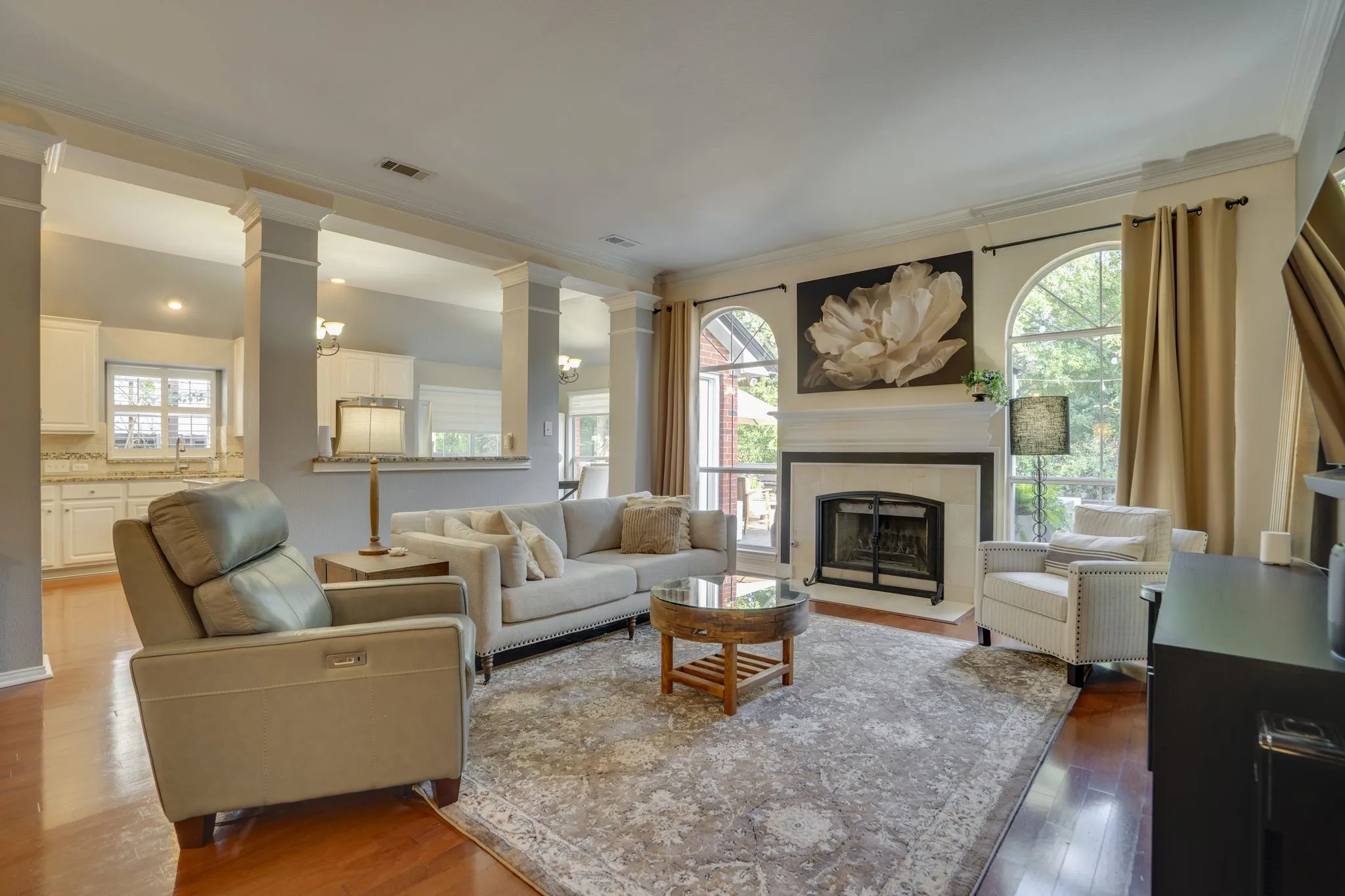 Hardwood flooring, fireplace with stone surround, crown molding, arched windows, and decorative columns