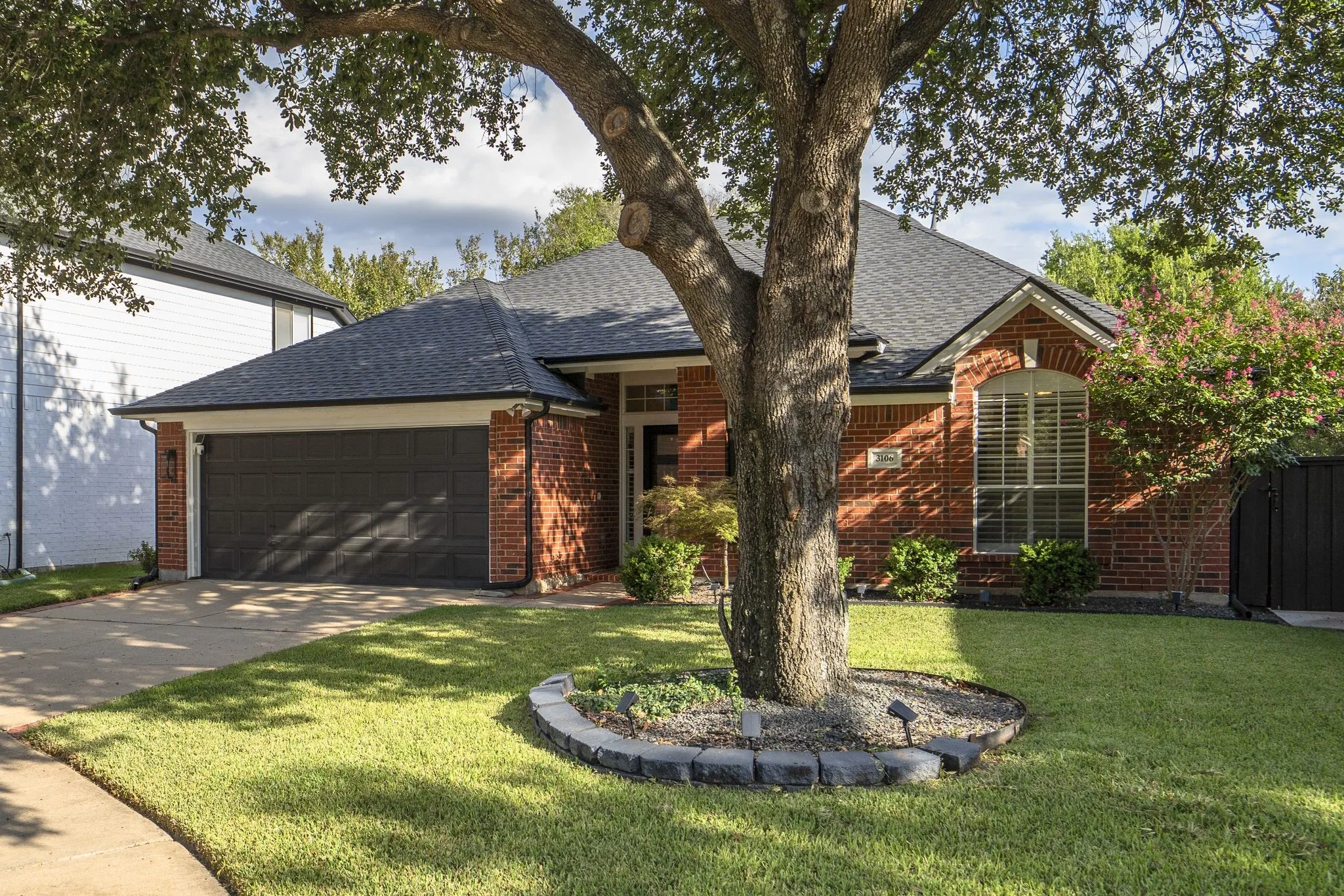 Brick exterior with an attached garage, featuring a concrete driveway and a manicured lawn with mature trees and shrubbery