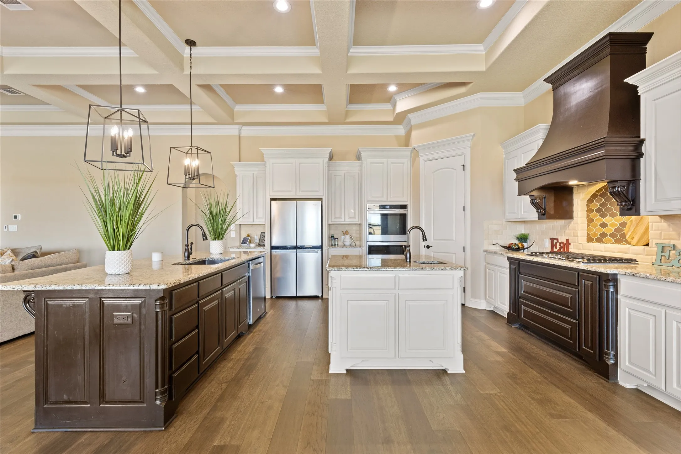 Kitchen featuring wood-finish flooring, a coffered ceiling, dual islands with undermount sinks, stainless steel appliances, and a decorative range hood