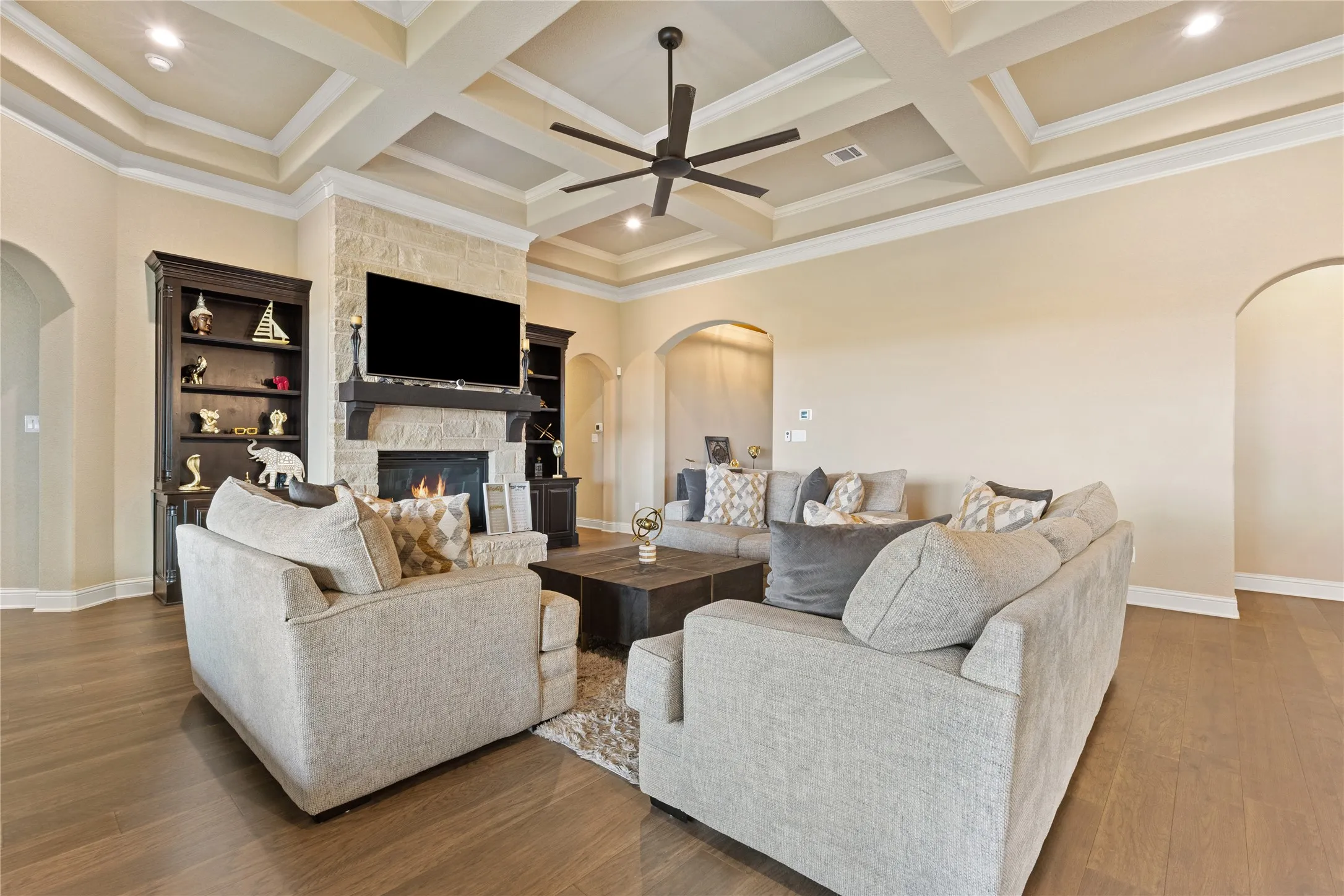 Coffered ceiling with a modern ceiling fan, stone-faced fireplace with a dark wood mantle, wood-finish flooring, and arched doorways