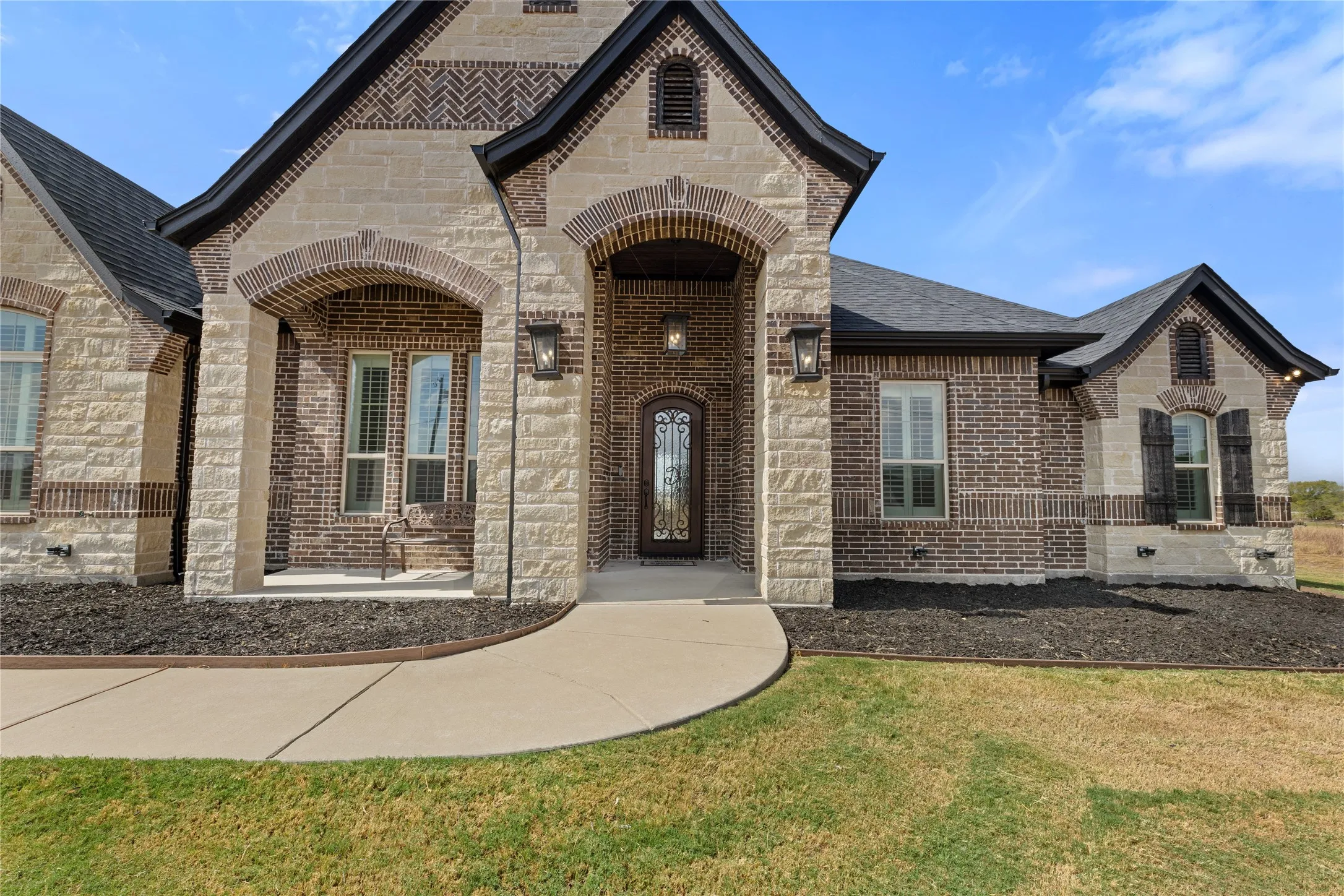 Stone and brick facade with arched entryways, dark shingle roof, decorative window shutters, and a custom front door with intricate glasswork