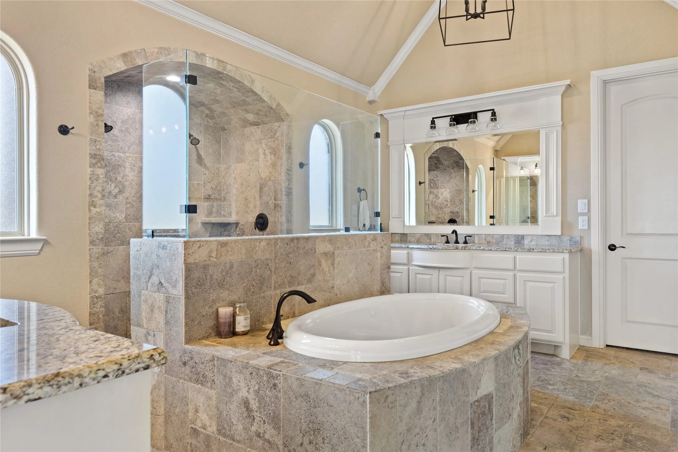 Built-in oval soaking tub, expansive tiled shower with glass enclosure, arched windows, dual vanity with undermount sinks, and a vaulted ceiling