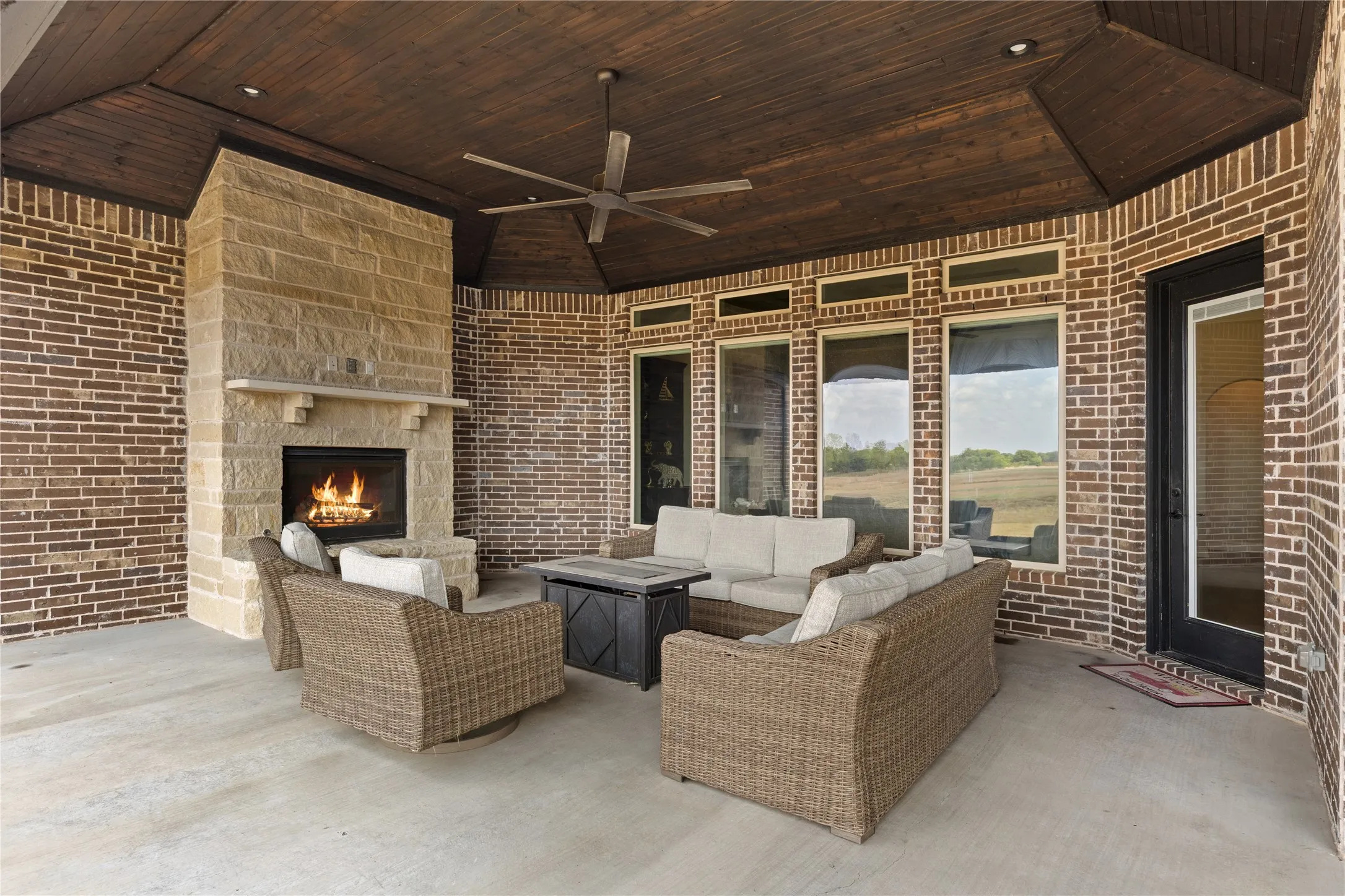 Covered patio featuring a stone fireplace, wood-plank ceiling, ceiling fan, brick exterior, and multiple tall windows