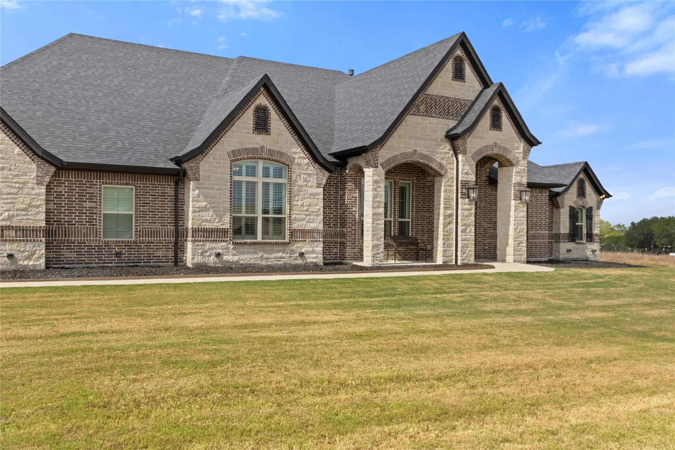 Stone and brick exterior with arched entryways, dark shingle roof, and exterior lighting fixtures