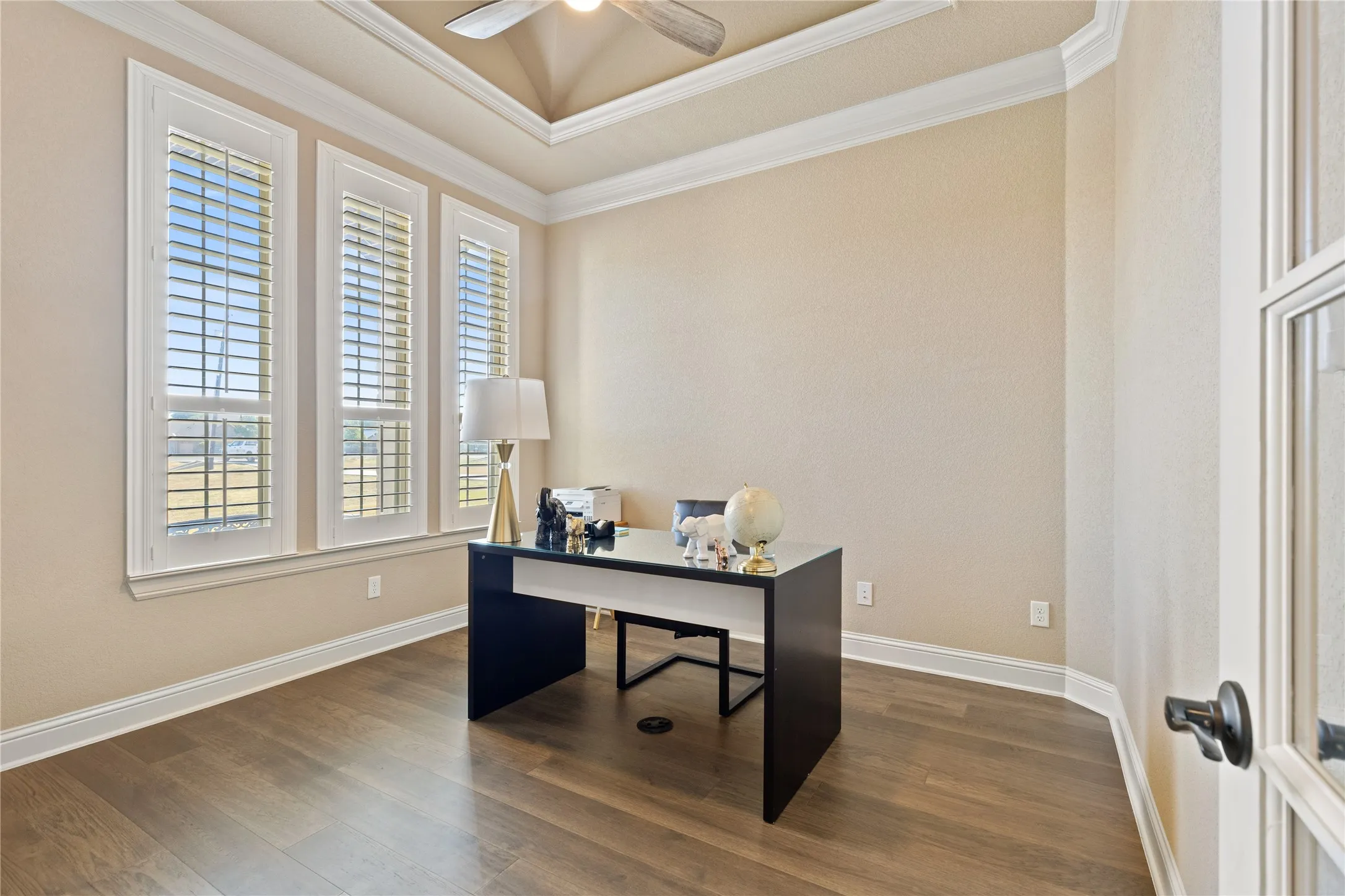 Tray ceiling with crown molding, wood-finish flooring, three windows with plantation shutters, and a ceiling fan