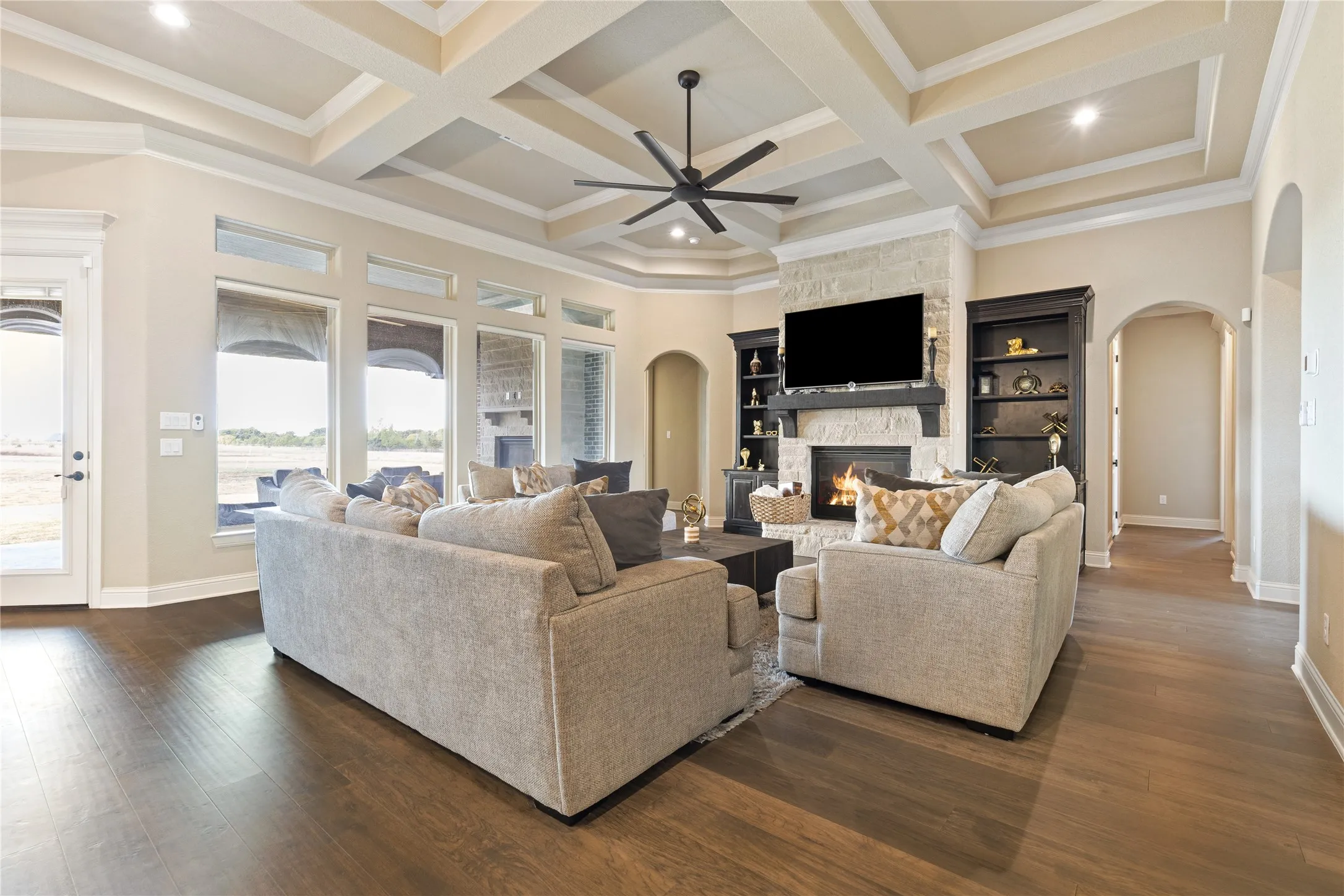 Coffered ceiling with recessed lighting and a ceiling fan, stone-faced fireplace with a dark wood mantel, built-in shelving, wood-finish flooring, and arched doorways