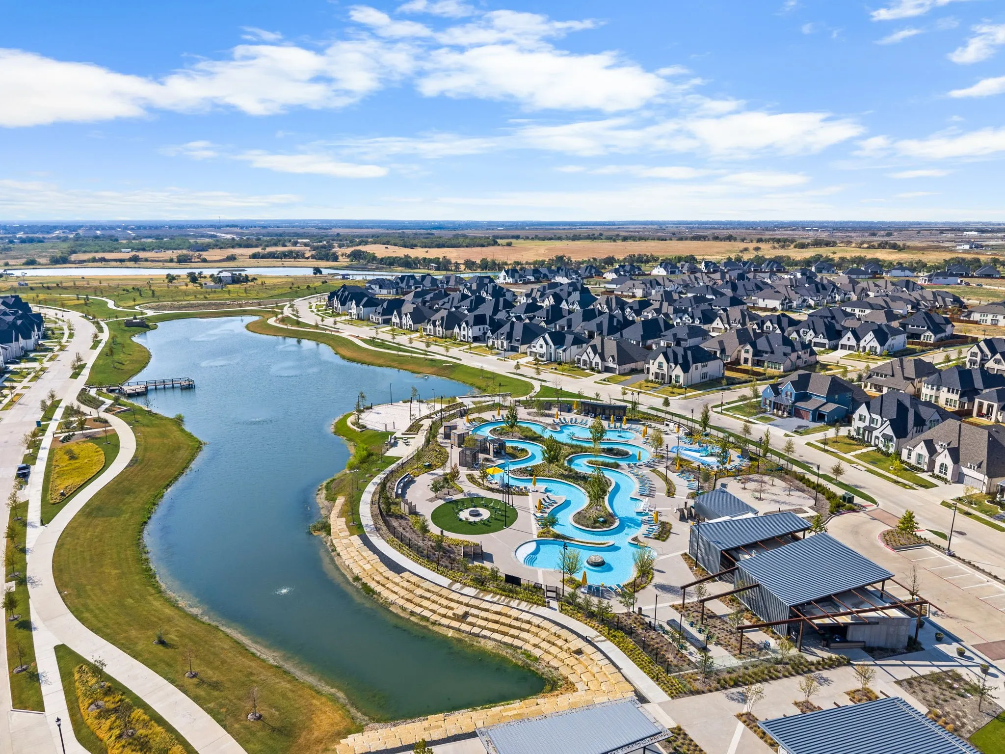 Expansive community lake with a dock, winding pathways, a water park with multiple pools, and covered gathering areas, all surrounded by residential properties